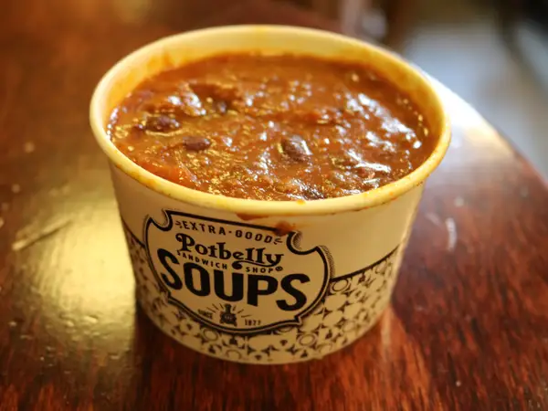 A paper cup labeled “Potbelly Soups” is filled with chunky chili, featuring beans and meat in a thick, reddish-brown sauce, placed on a wooden surface.