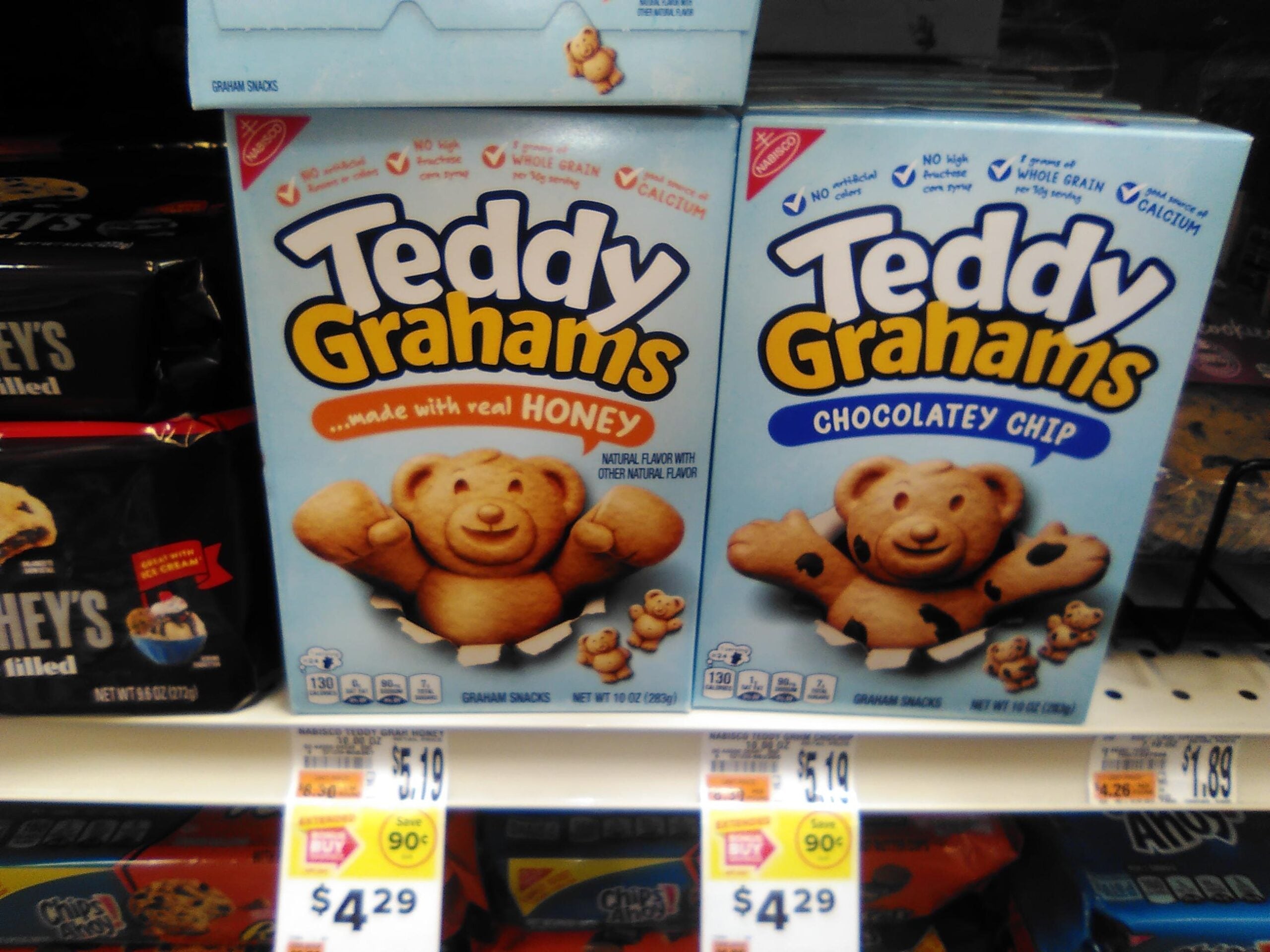 Boxes of Teddy Grahams snacks—Honey and Chocolate Chip flavors—are displayed on a grocery store shelf, each box featuring a smiling bear-shaped graham cracker. Price tags are visible below the boxes.