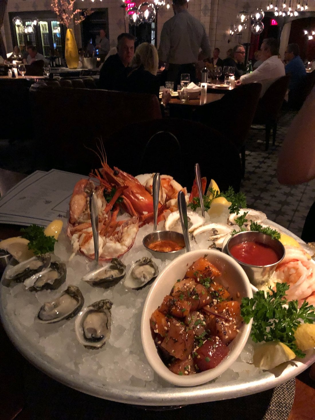A seafood platter on ice with oysters, shrimp, poke, lemon wedges, parsley, and dipping sauces, served at a dimly lit restaurant with diners in the background.