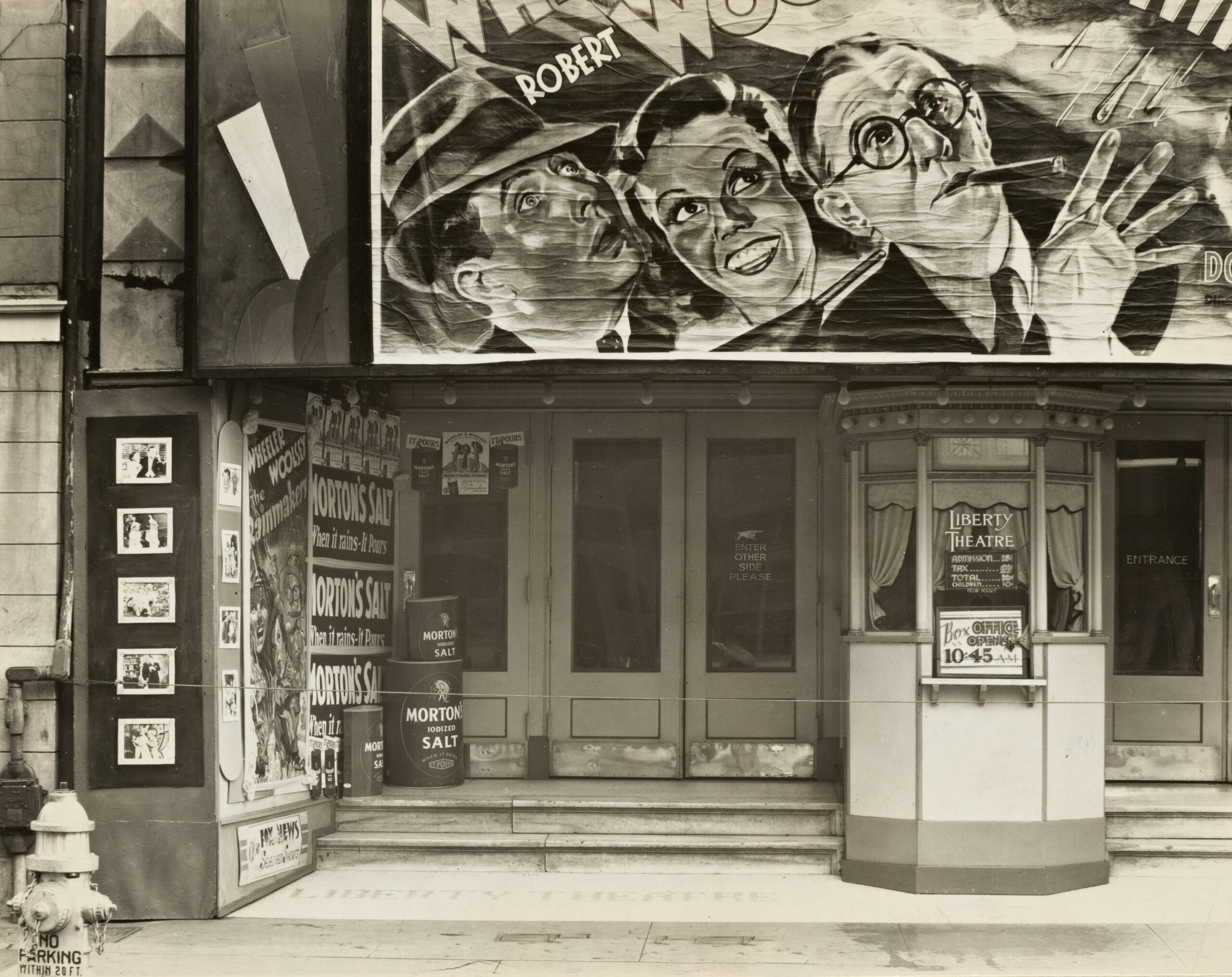 Black-and-white photo of a theater entrance with movie posters and a large illustrated banner featuring three faces above the doors. A display case shows showtimes, and a fire hydrant is on the sidewalk.