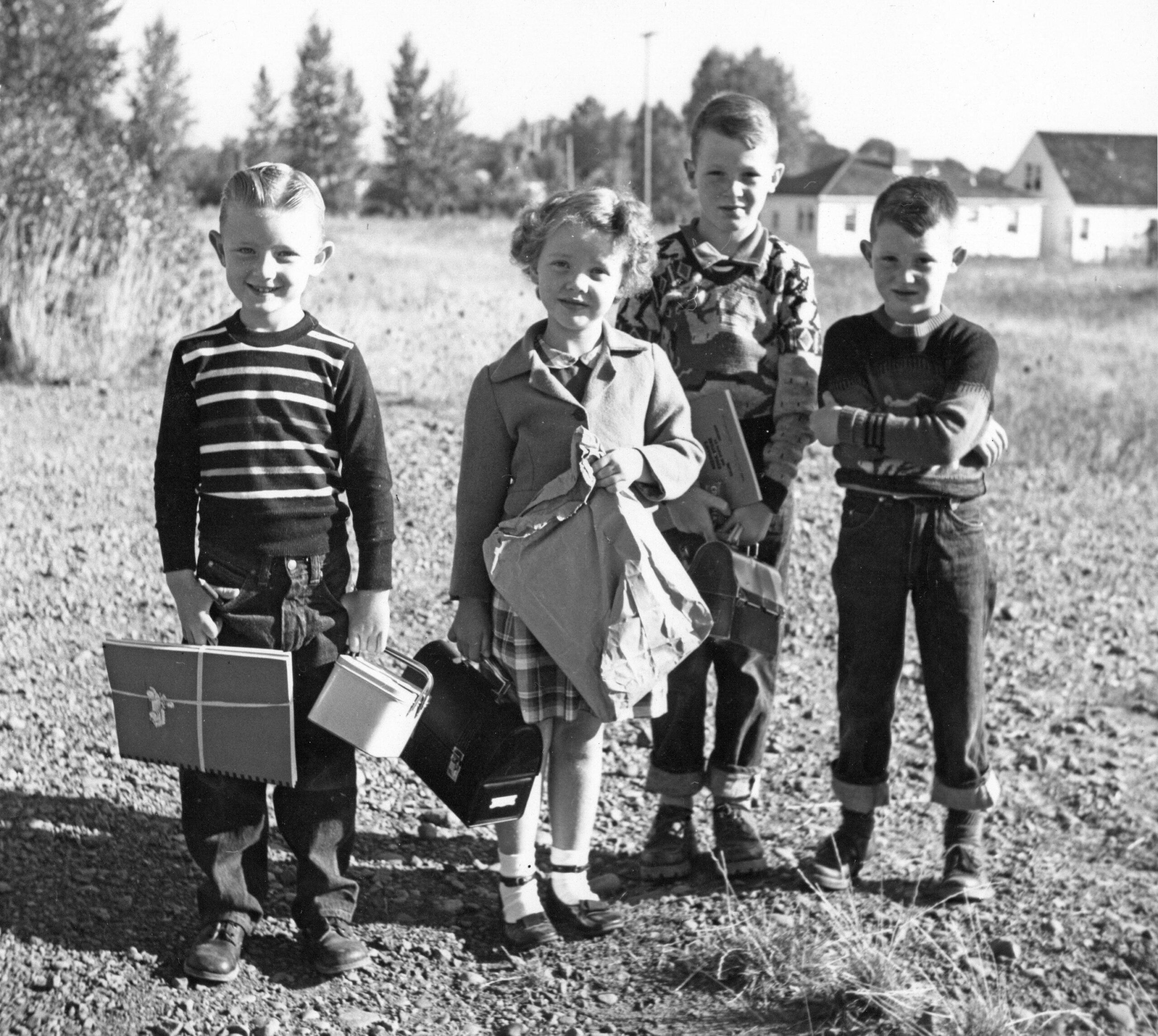 Four children stand outdoors on a dirt path, holding lunchboxes and bags. Trees and houses are visible in the background. The photo appears to be from an earlier decade, possibly the 1950s or 1960s.