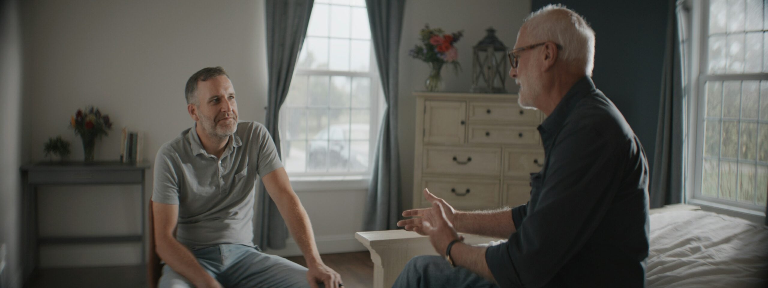 Two men sit facing each other in a softly lit bedroom, engaged in conversation about boomers saving money. One gestures with his hands while the other listens, both appearing relaxed. Large windows and light-colored furniture are visible in the background.