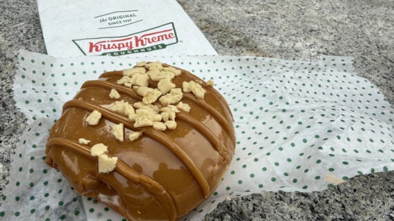 A Krispy Kreme doughnut with caramel glaze and cookie crumbles sits on a dotted paper, with a Krispy Kreme napkin in the background on a gray stone surface.