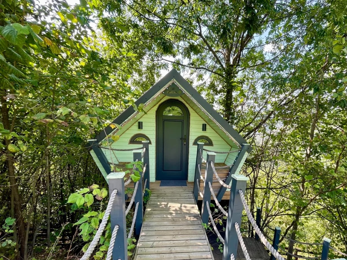 A small green cabin with arched windows and a black door sits among dense trees, accessed by a wooden rope bridge.