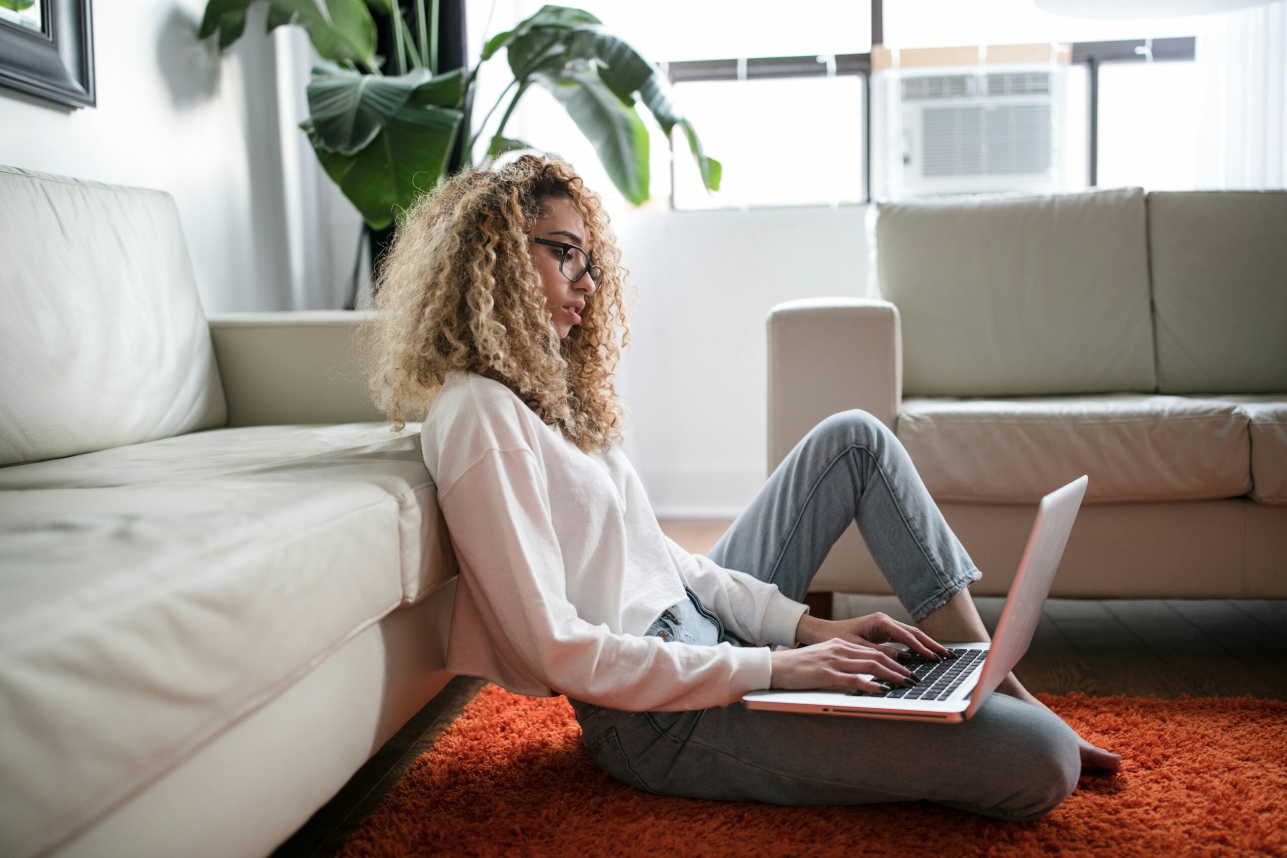 A person with curly hair and glasses sits on an orange rug, leaning against a white sofa, working on a laptop in a bright living room with plants and large windows—an everyday scene that perfectly captures life in 2026.