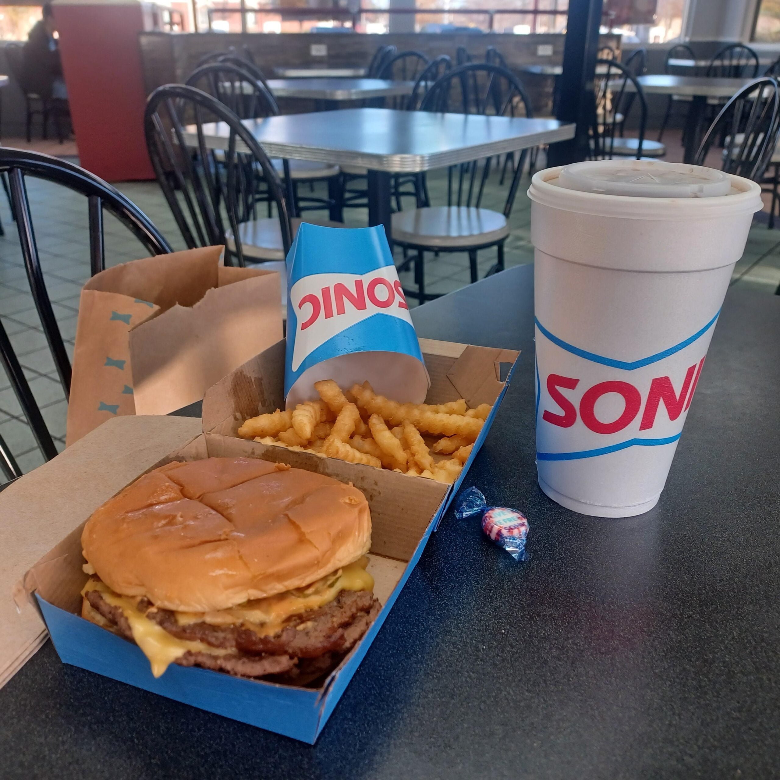 A Sonic meal with a cheeseburger, crinkle-cut fries, a large drink cup, a paper bag, and two wrapped peppermint candies on a table in a fast-food restaurant.