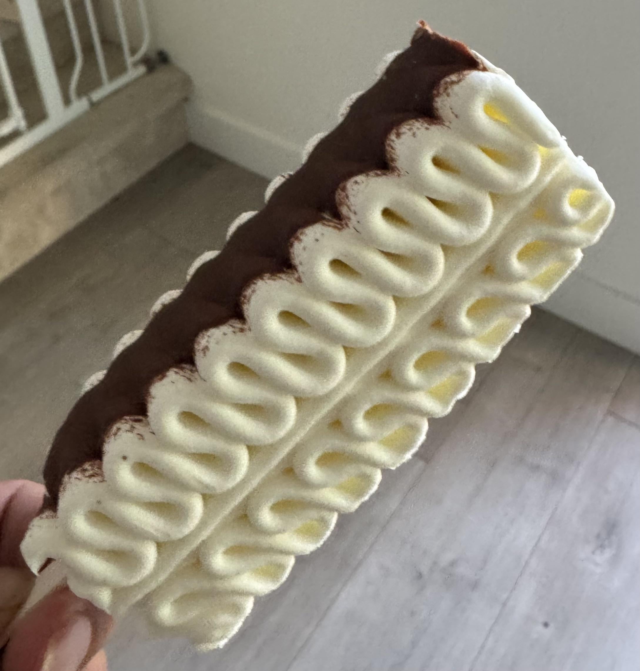 A hand holds an ice cream sandwich with wavy white ice cream layers and a chocolate layer, indoors on a wood floor background.