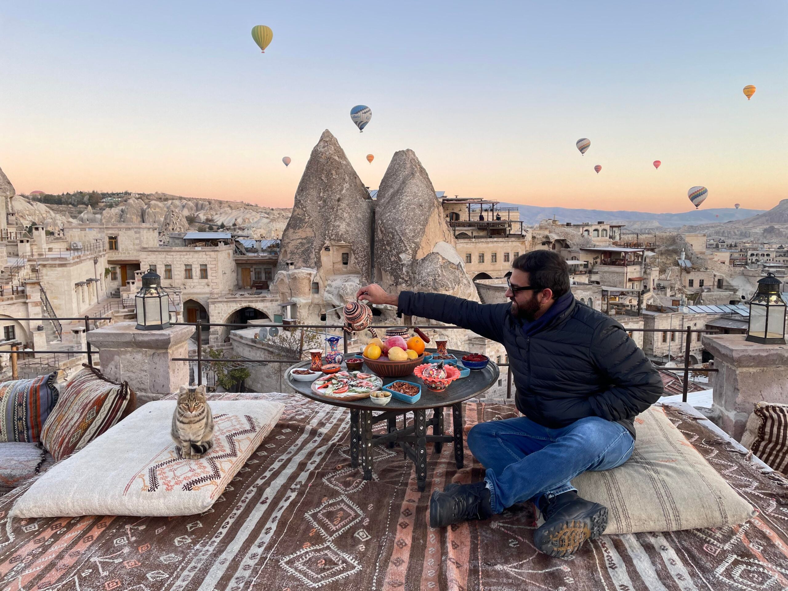 A man pours tea at a rooftop breakfast with various dishes. Hot air balloons float over a unique rocky landscape at sunrise. A cat sits nearby on a cushion.