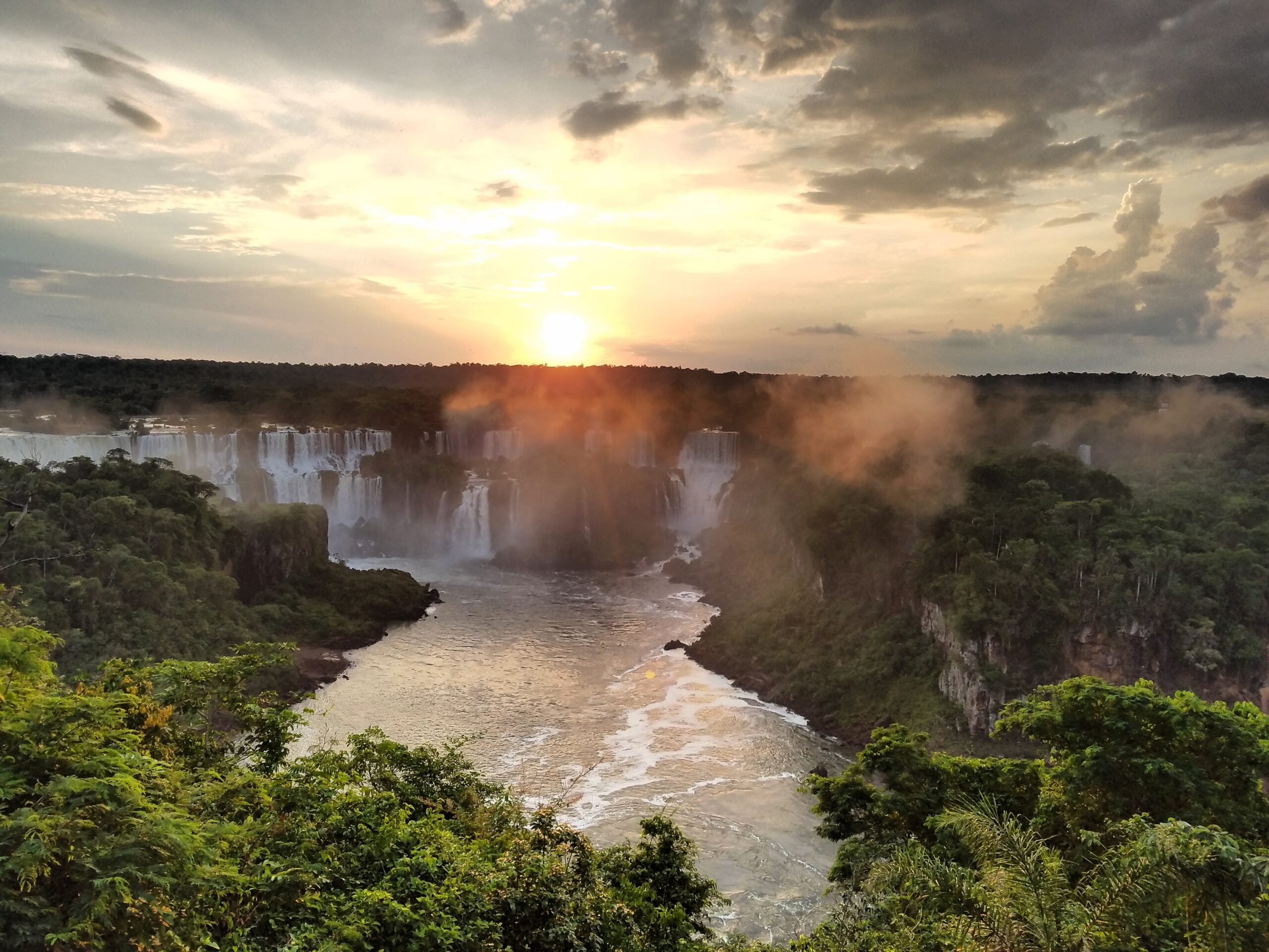 Sunset over Iguazu Falls, with mist rising from the waterfalls, lush green forest surrounding the falls, and a cloudy sky illuminated by the setting sun.