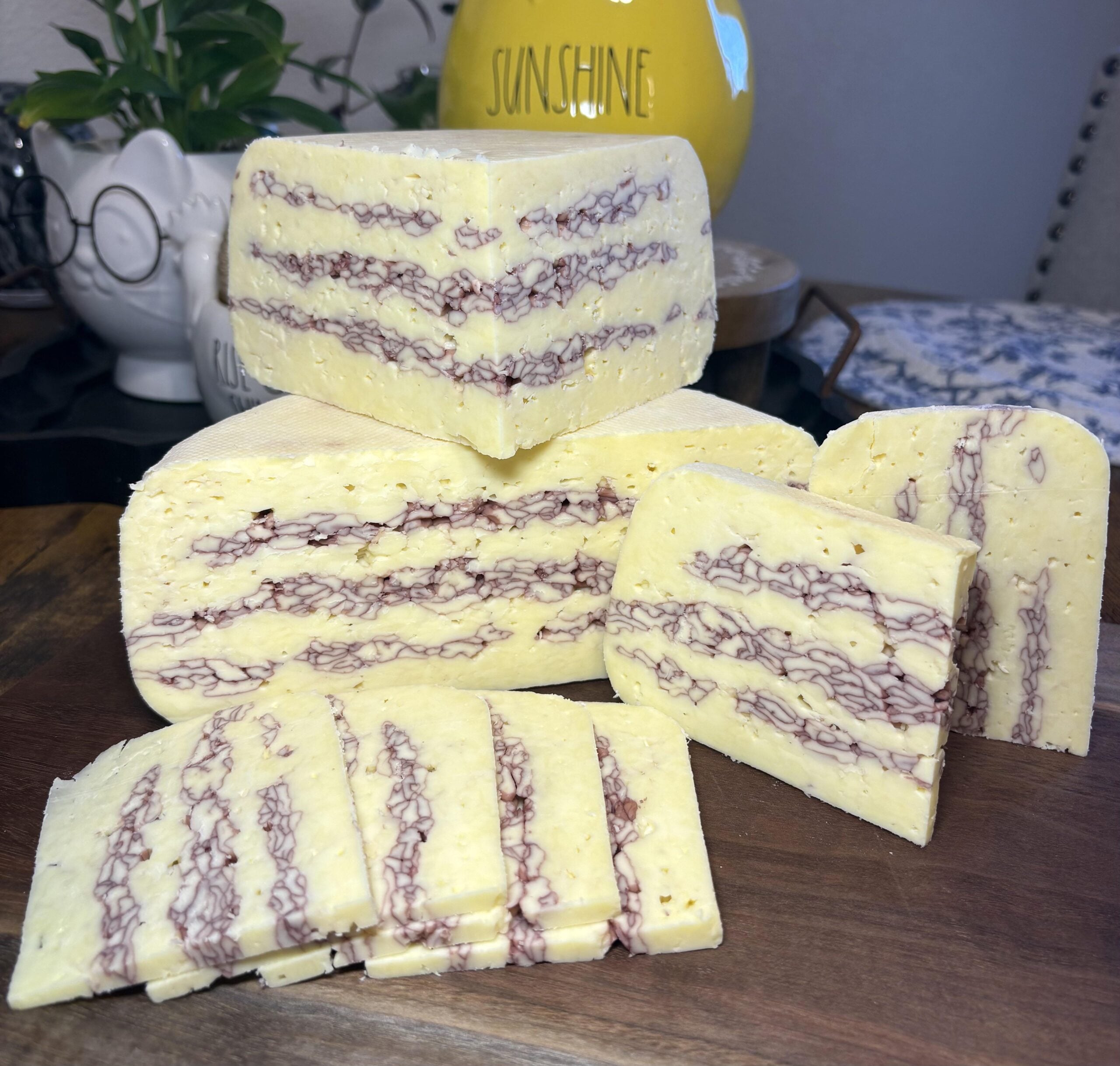 Several blocks and slices of white cheese with distinctive purple marbling are arranged on a wooden board, with a yellow jar labeled “SUNSHINE” and a plant in the background.