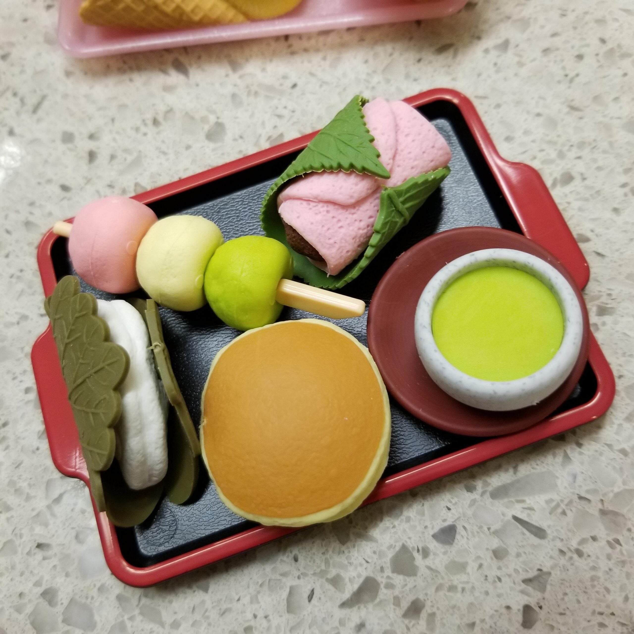 A tray with Japanese sweets: dango skewers, sakura mochi, a pancake sandwich, a leaf-wrapped sweet, and a bowl of green matcha tea on a speckled countertop.
