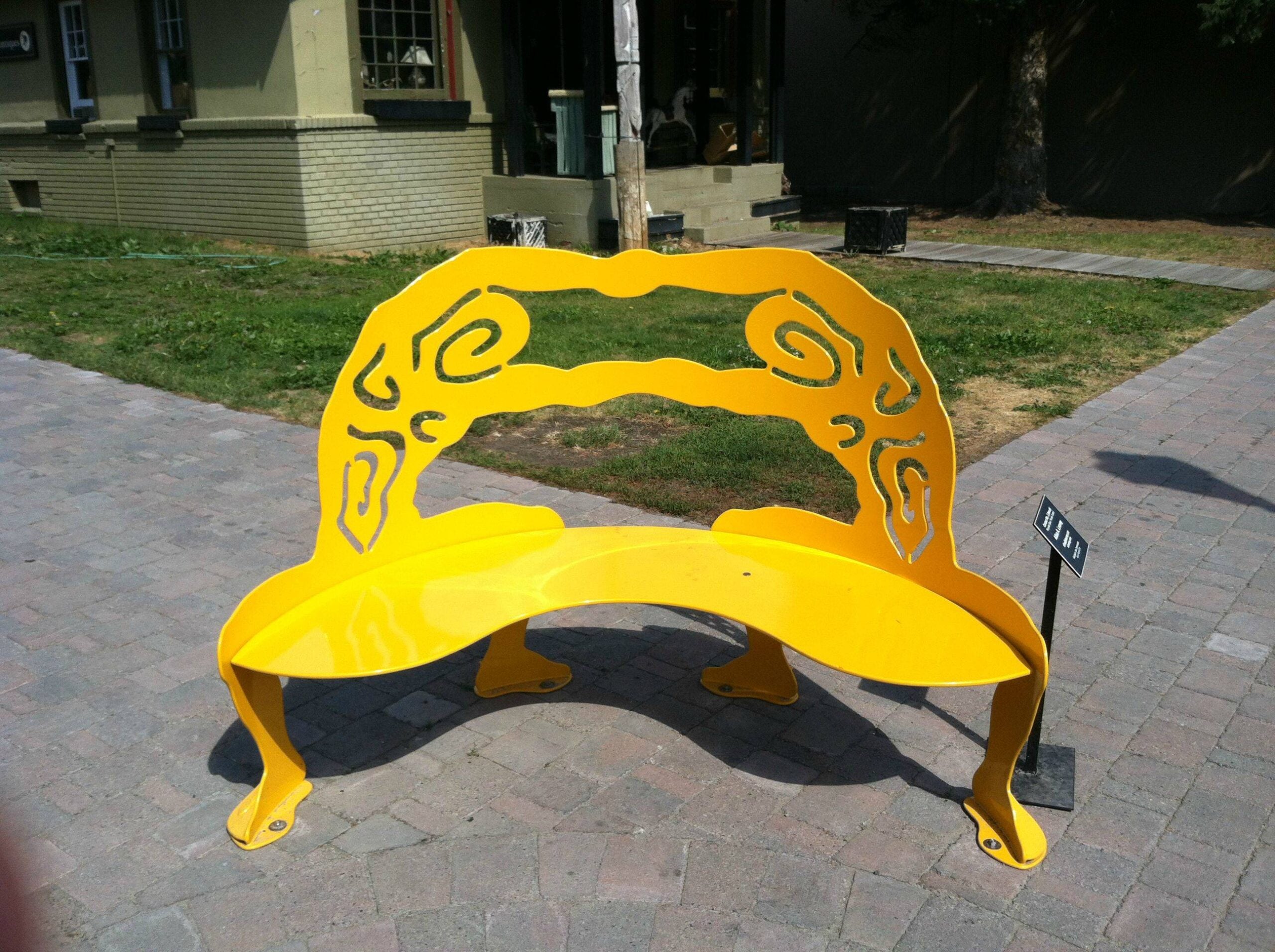 A bright yellow, artistic metal bench with decorative swirls on the backrest sits on a stone walkway in an outdoor urban area.