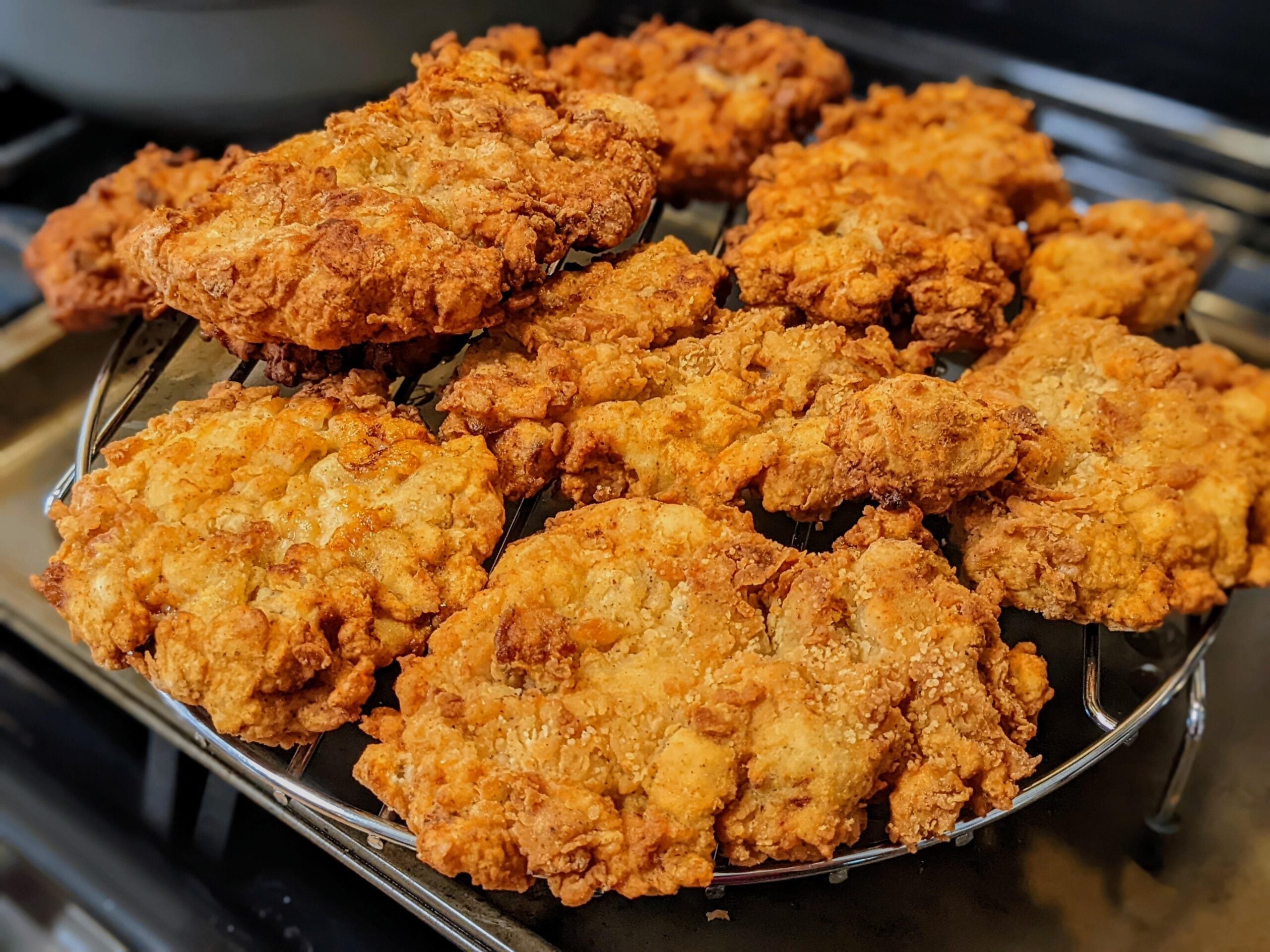 Golden brown, crispy fried chicken pieces cooling on a wire rack over a stovetop, with crunchy textured breading visible on each piece.