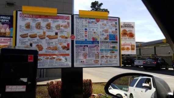 A drive-thru menu at a fast food restaurant, showing various meal options, including burgers, sandwiches, chicken, and drinks. Cars are visible in the background and a side mirror reflects part of a white vehicle.