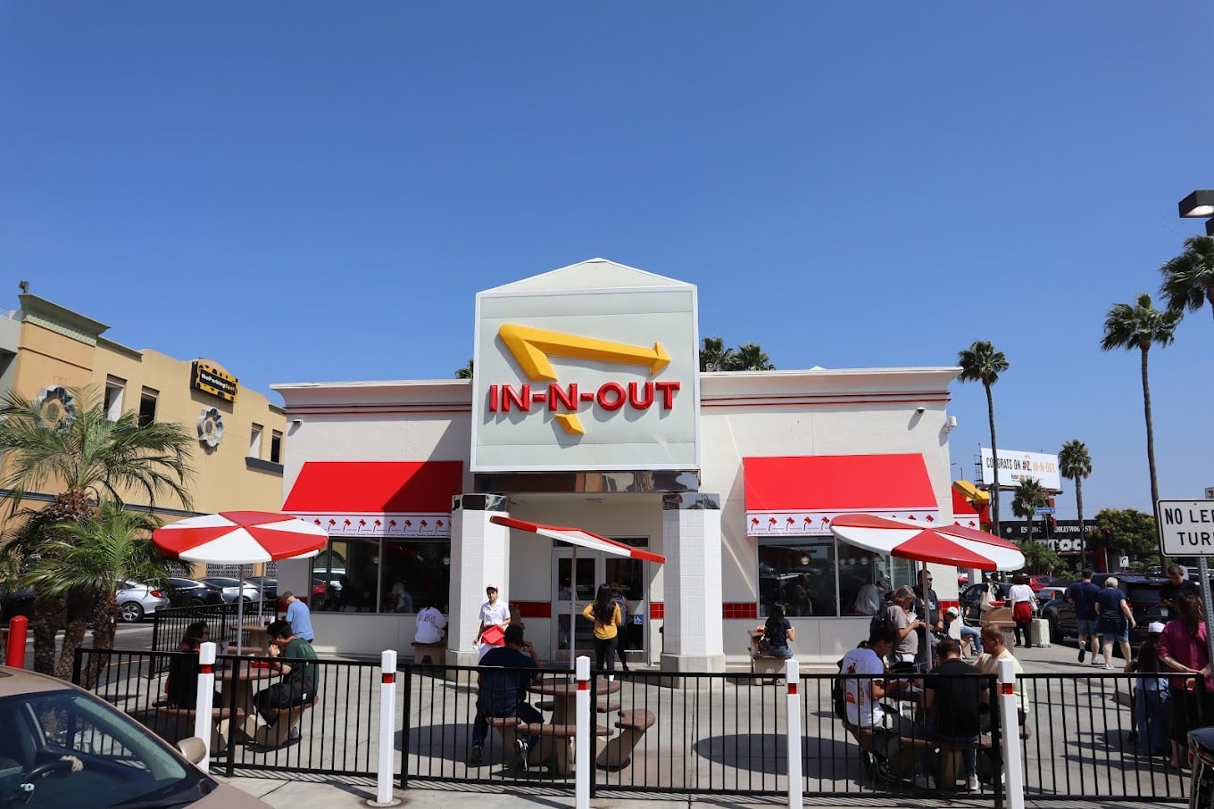 People sit under red-and-white umbrellas outside an In-N-Out Burger restaurant with its red, yellow, and white signage, on a sunny day with palm trees in the background.