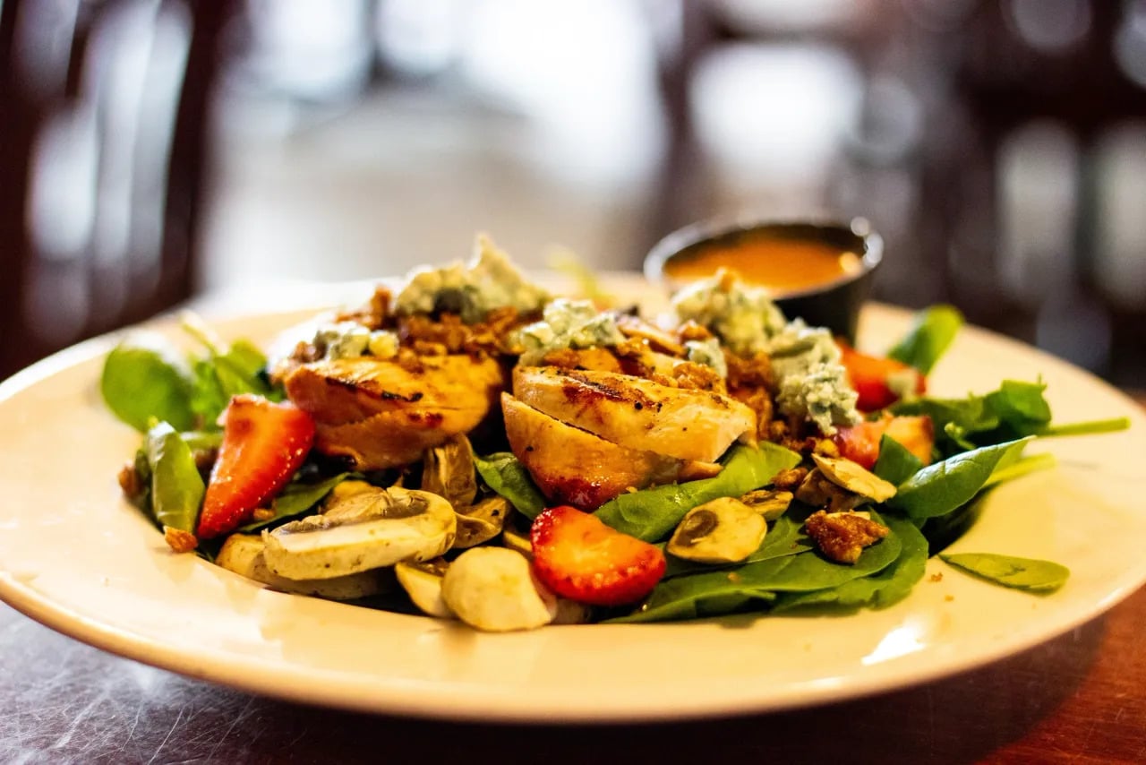 A salad with grilled chicken, spinach, strawberries, mushrooms, blue cheese, and nuts, served on a white plate with a small cup of dressing in the background.