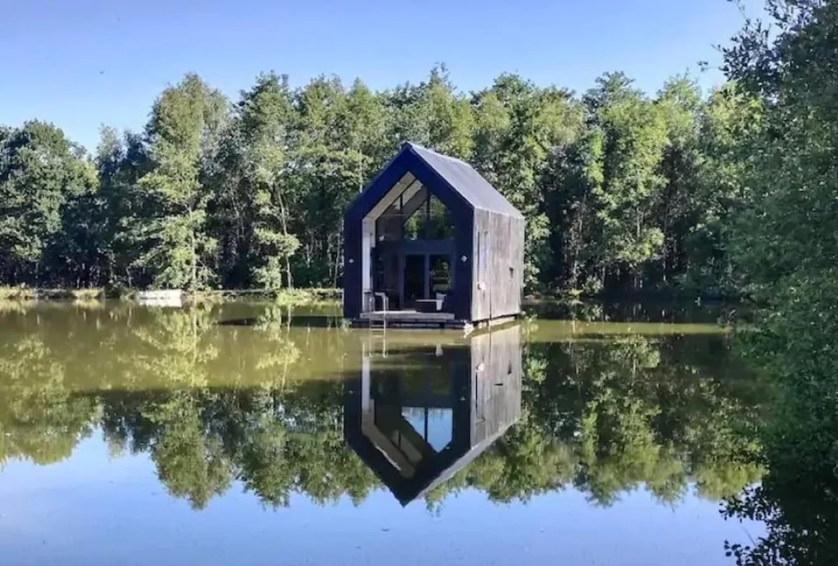 A small, modern black cabin sits on a calm lake, surrounded by lush green trees. The cabin and trees are reflected clearly in the still water under a bright blue sky.