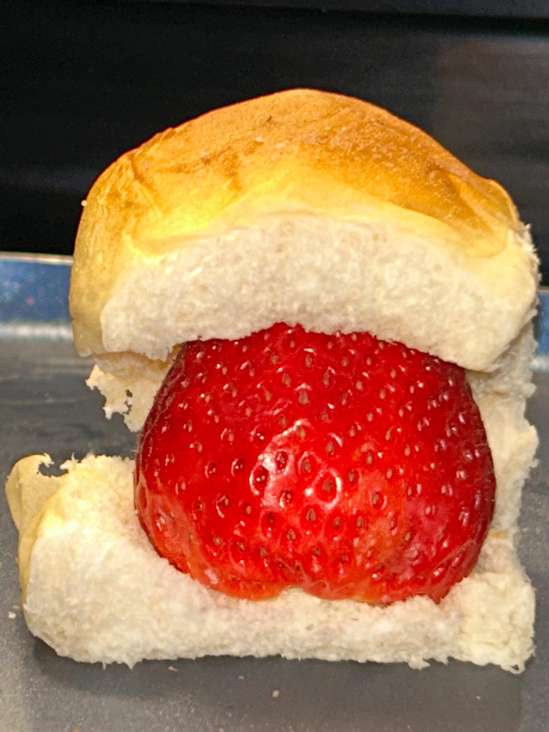 A close-up of a large, ripe strawberry sandwiched between two halves of a soft, golden-brown bread roll, resembling a strawberry burger.