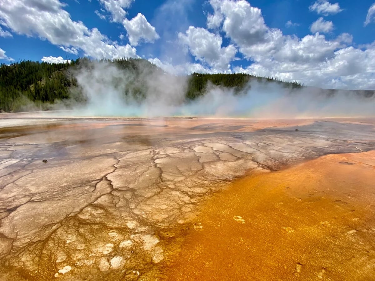 Colorful geothermal hot spring with orange and brown mineral deposits in the foreground, rising steam, and a forested hill under a blue sky with white clouds in the background.