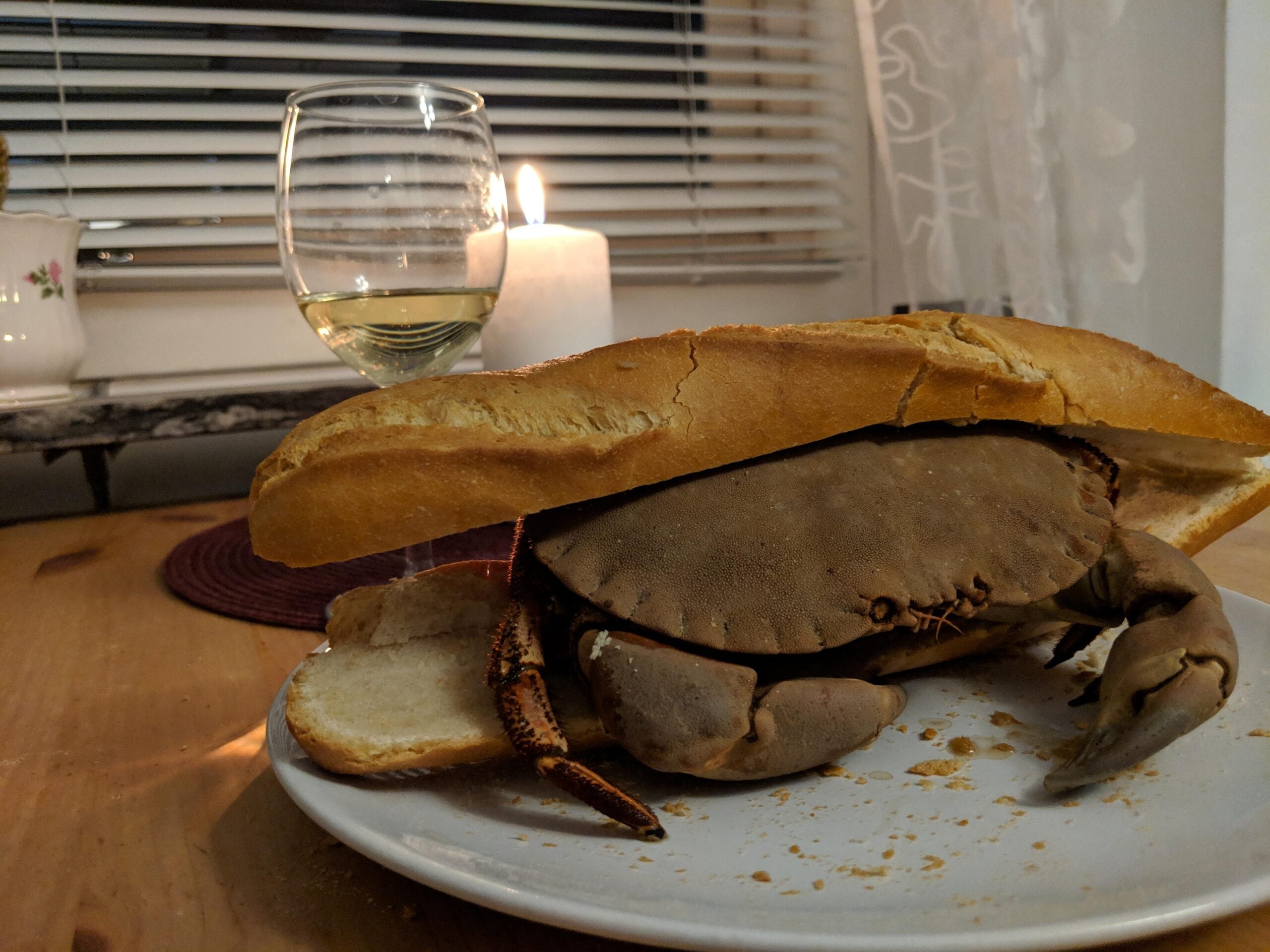 A whole crab placed inside a sliced baguette on a plate, resembling a sandwich. A glass of white wine and a lit candle sit in the background on a wooden table.