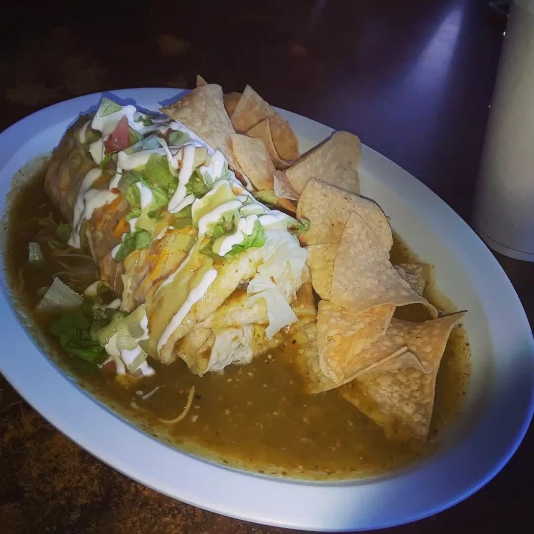 A large burrito topped with lettuce, cheese, sour cream, and diced tomatoes sits in green sauce, served with tortilla chips on a white plate.