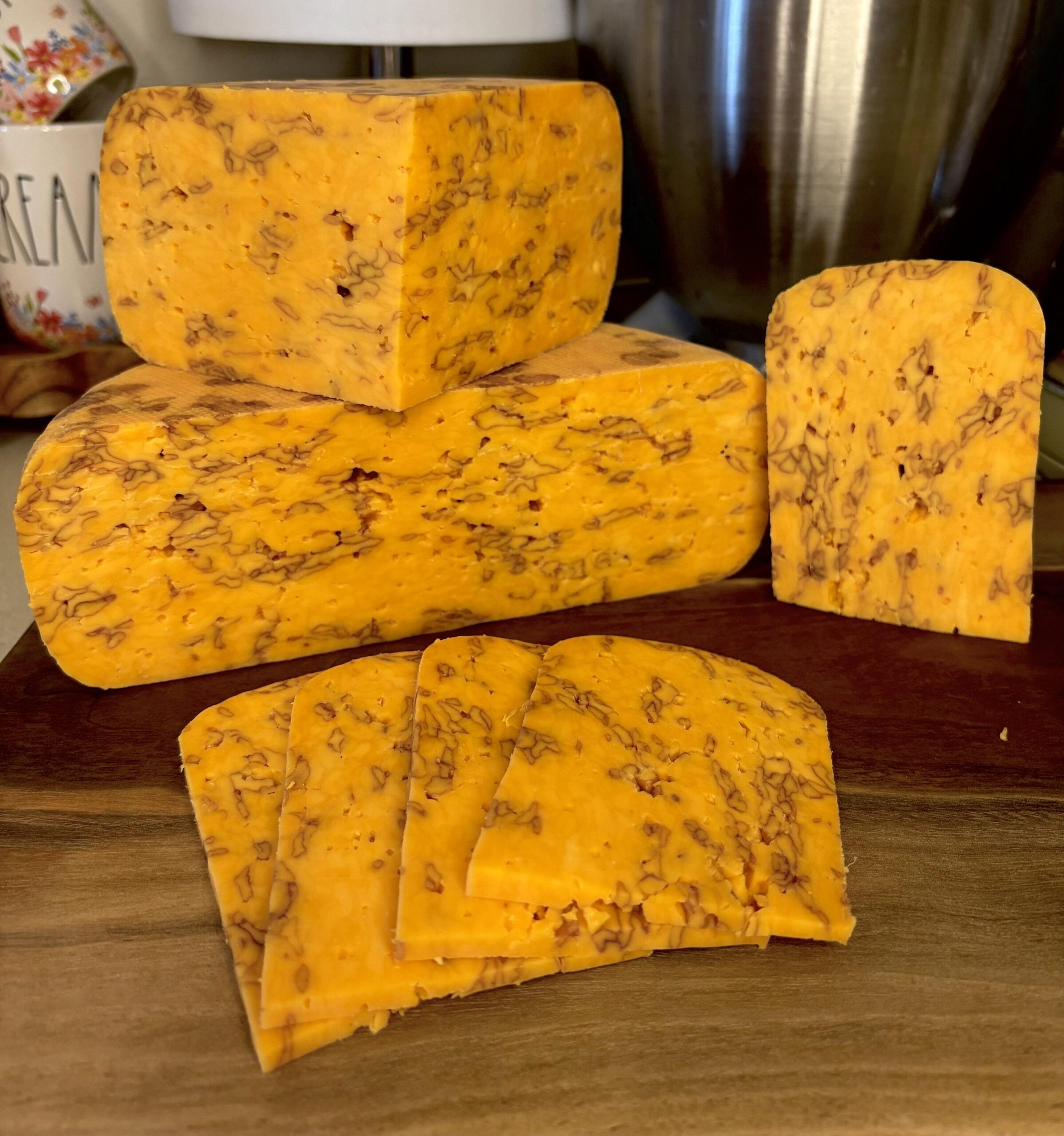 Several blocks and slices of orange marbled cheese are displayed on a wooden cutting board, with a kitchen setting visible in the background.