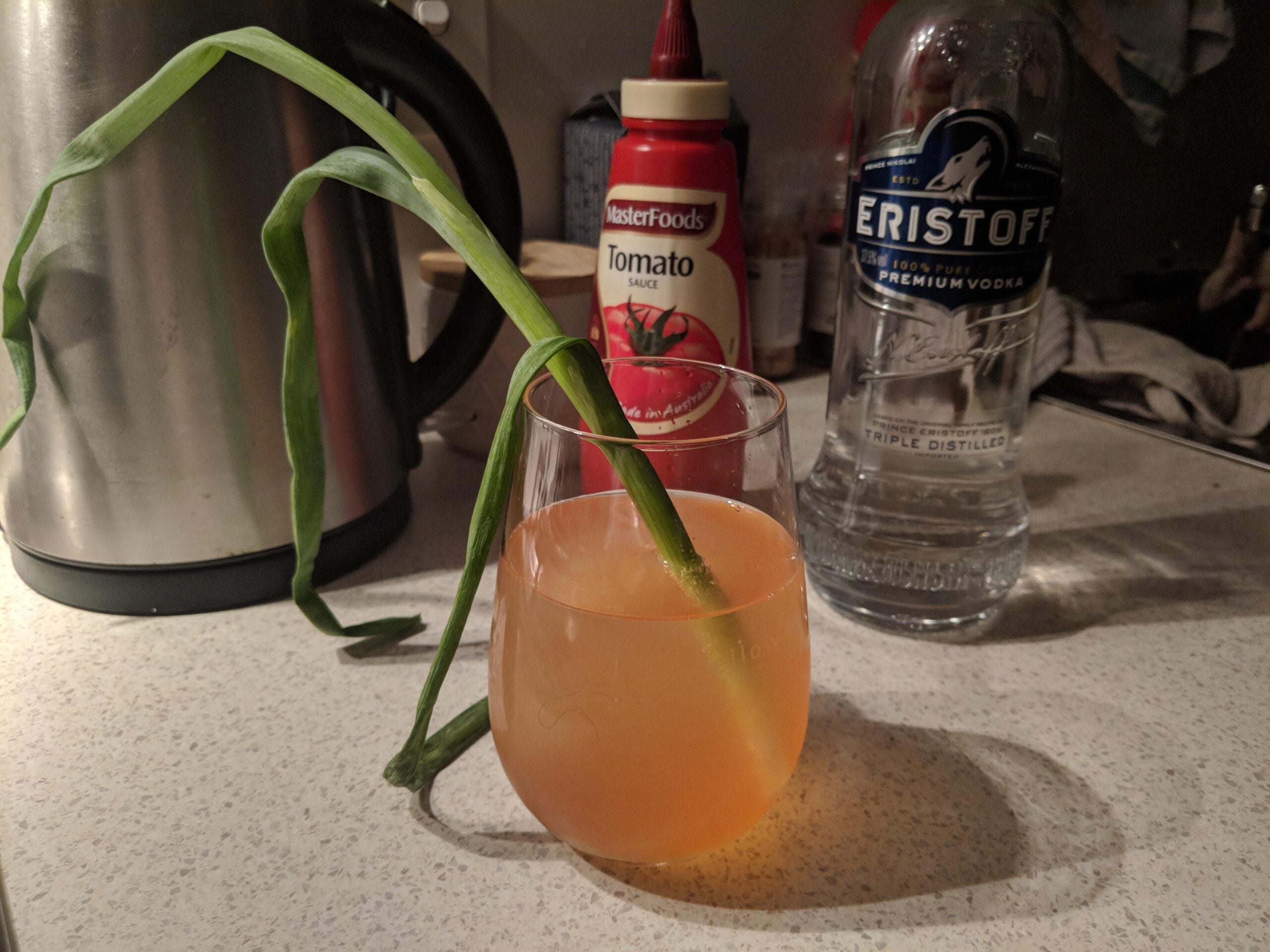A drink in a glass with a long green onion as a garnish, next to a bottle of vodka, tomato sauce, and a kettle on a kitchen counter.