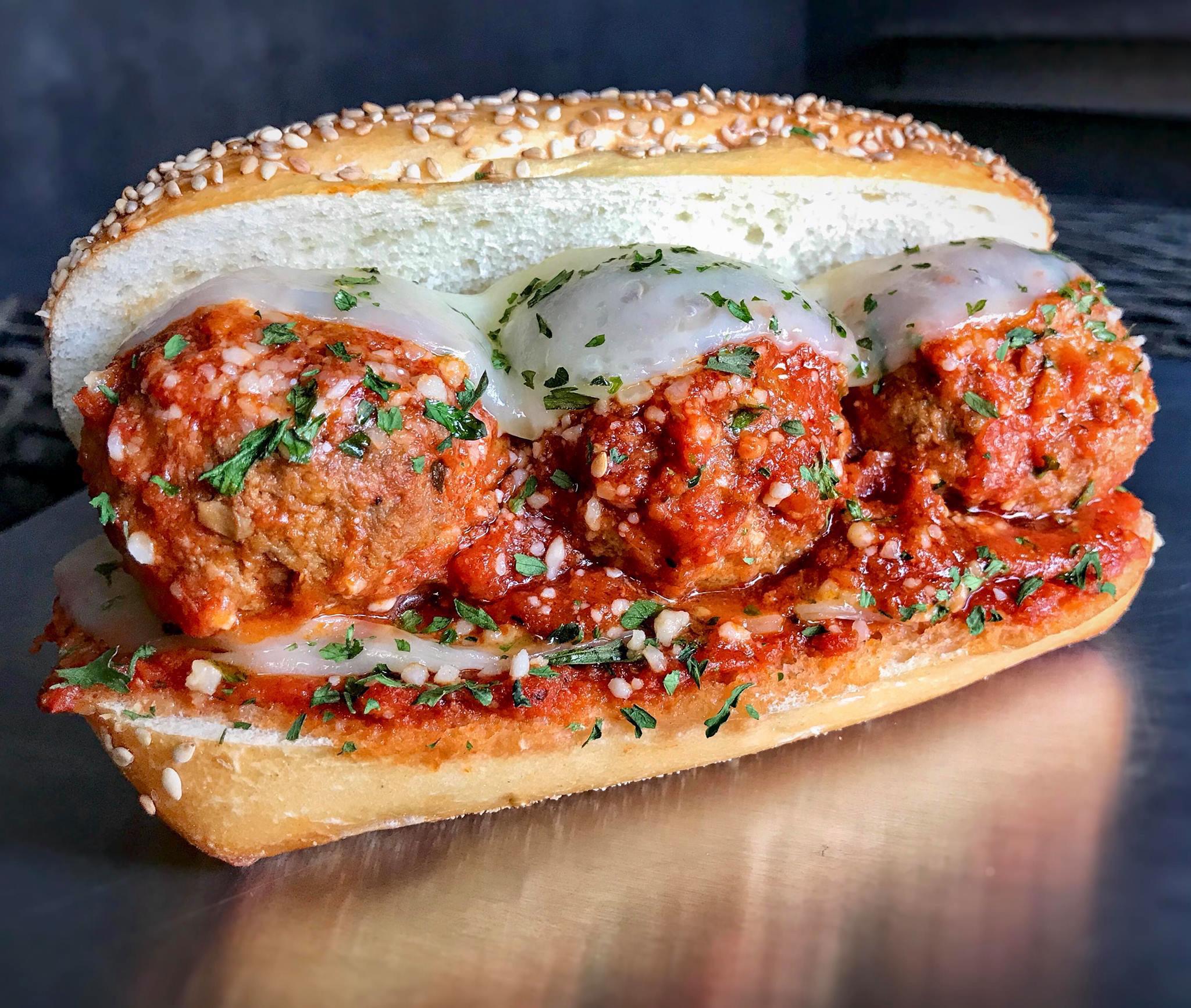 A sesame sub roll filled with three meatballs in marinara sauce, topped with melted cheese and chopped herbs, sits on a metallic surface.