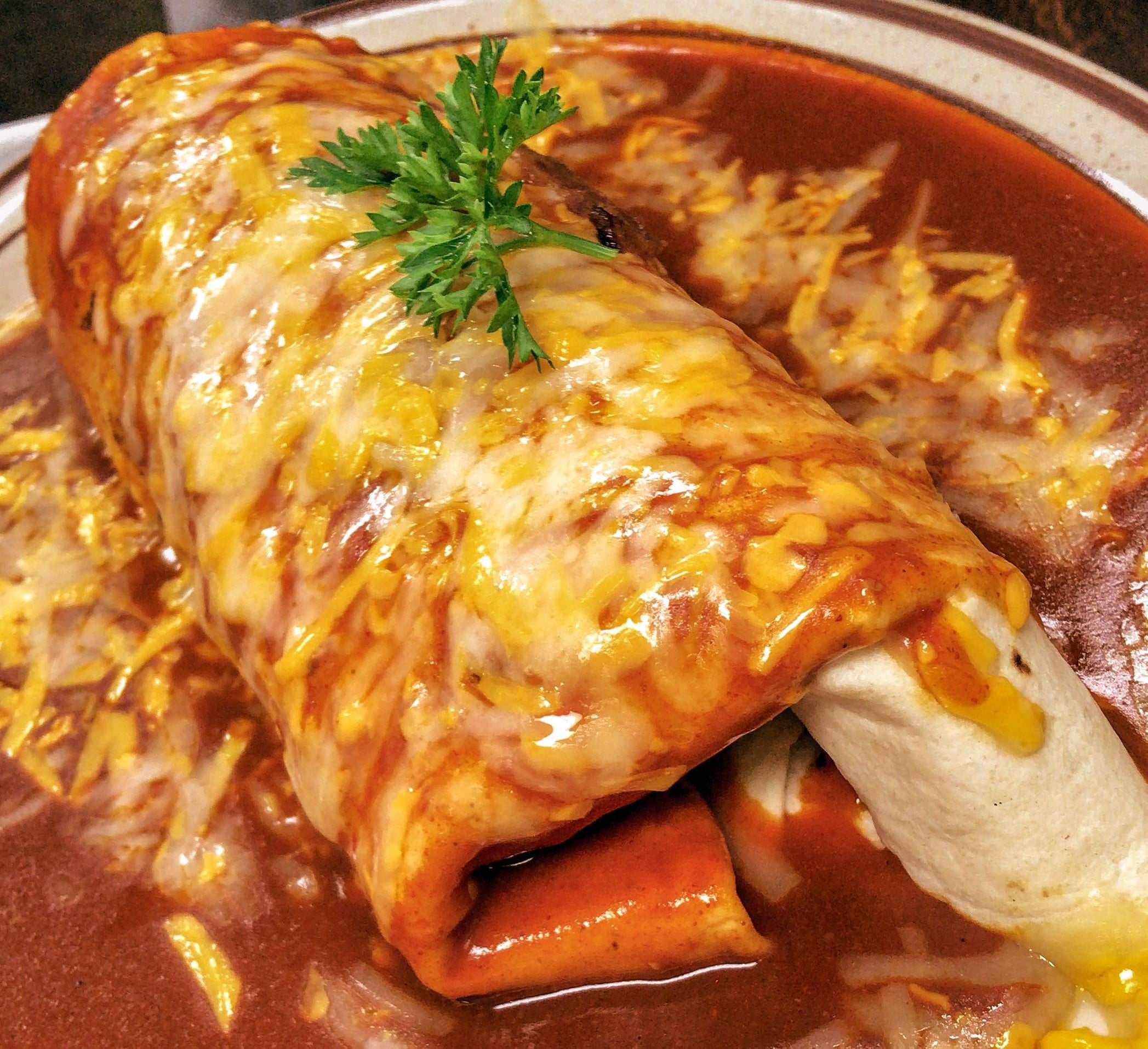 A large burrito topped with melted cheese and red enchilada sauce, garnished with parsley, sits on a plate with extra sauce and shredded cheese around it.