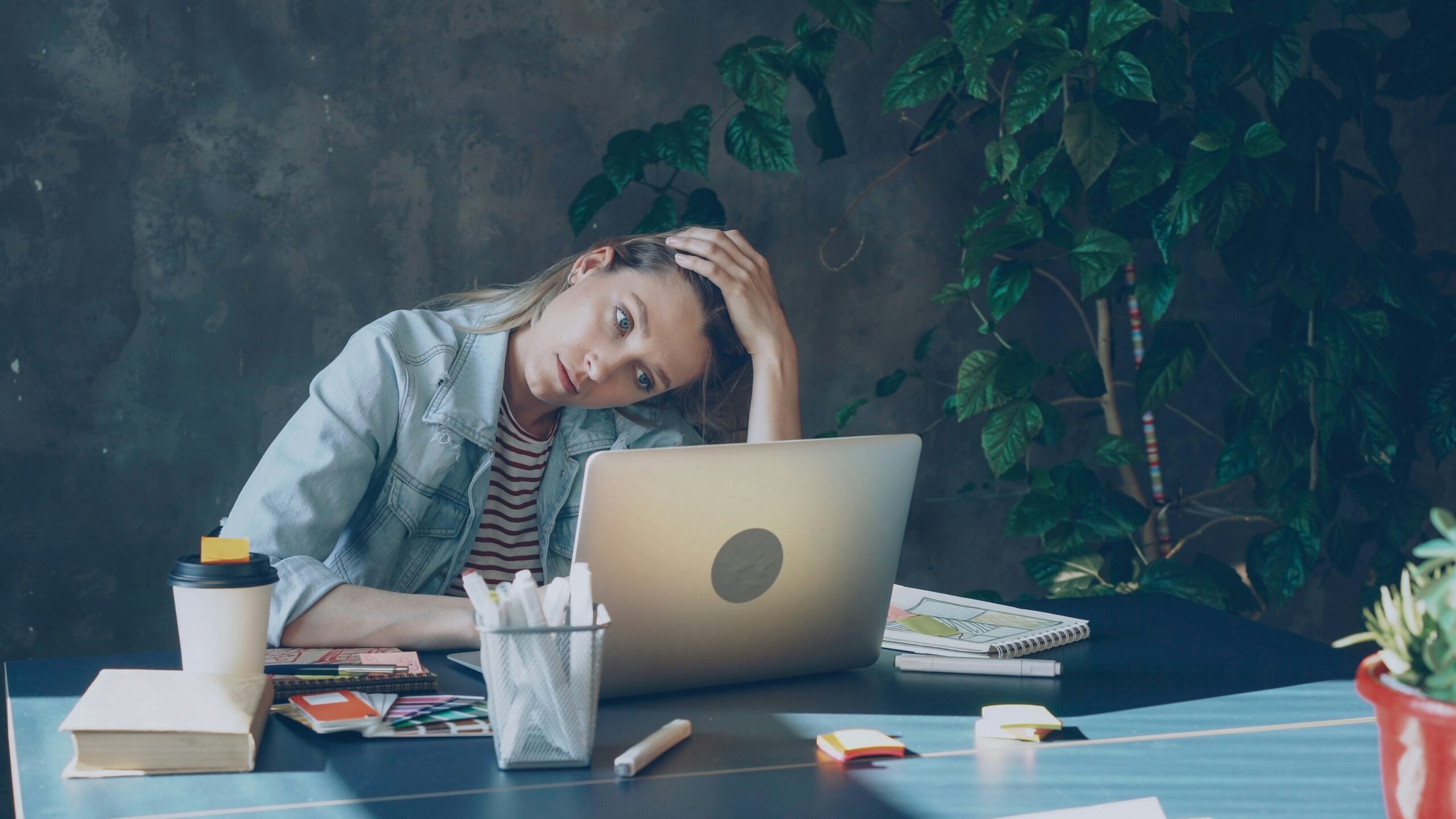 A person sits at a desk with a laptop, holding their head and looking tired—a familiar scene in life in 2026. The desk is scattered with notebooks, pens, a coffee cup, and post-it notes, while a green plant adds a touch of calm in the background.