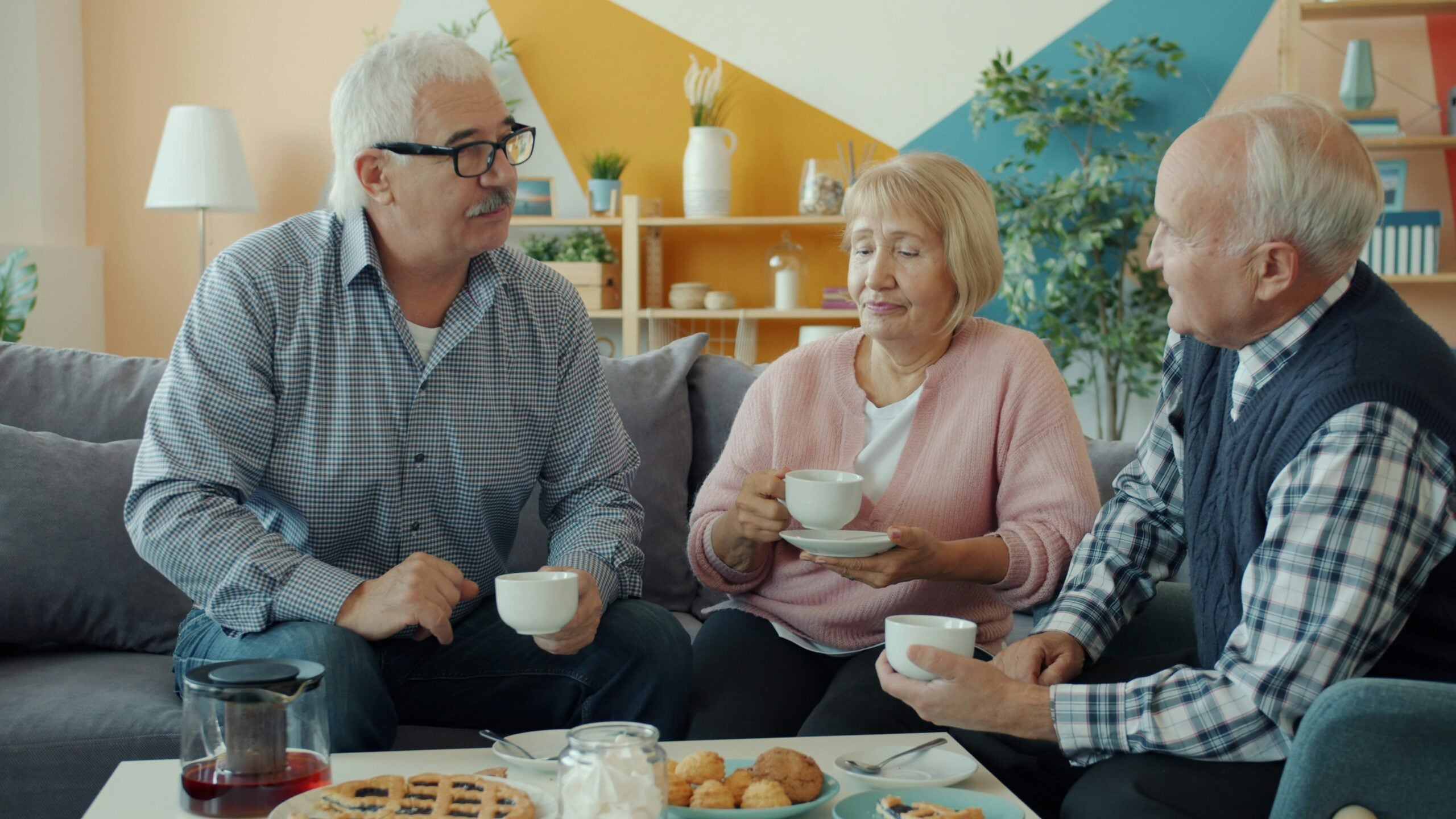 Four older adults sit on a couch, smiling and enjoying tea with pastries in a bright, cozy living room with plants and modern decor—proof that boomers saving money can still enjoy life’s little luxuries together.