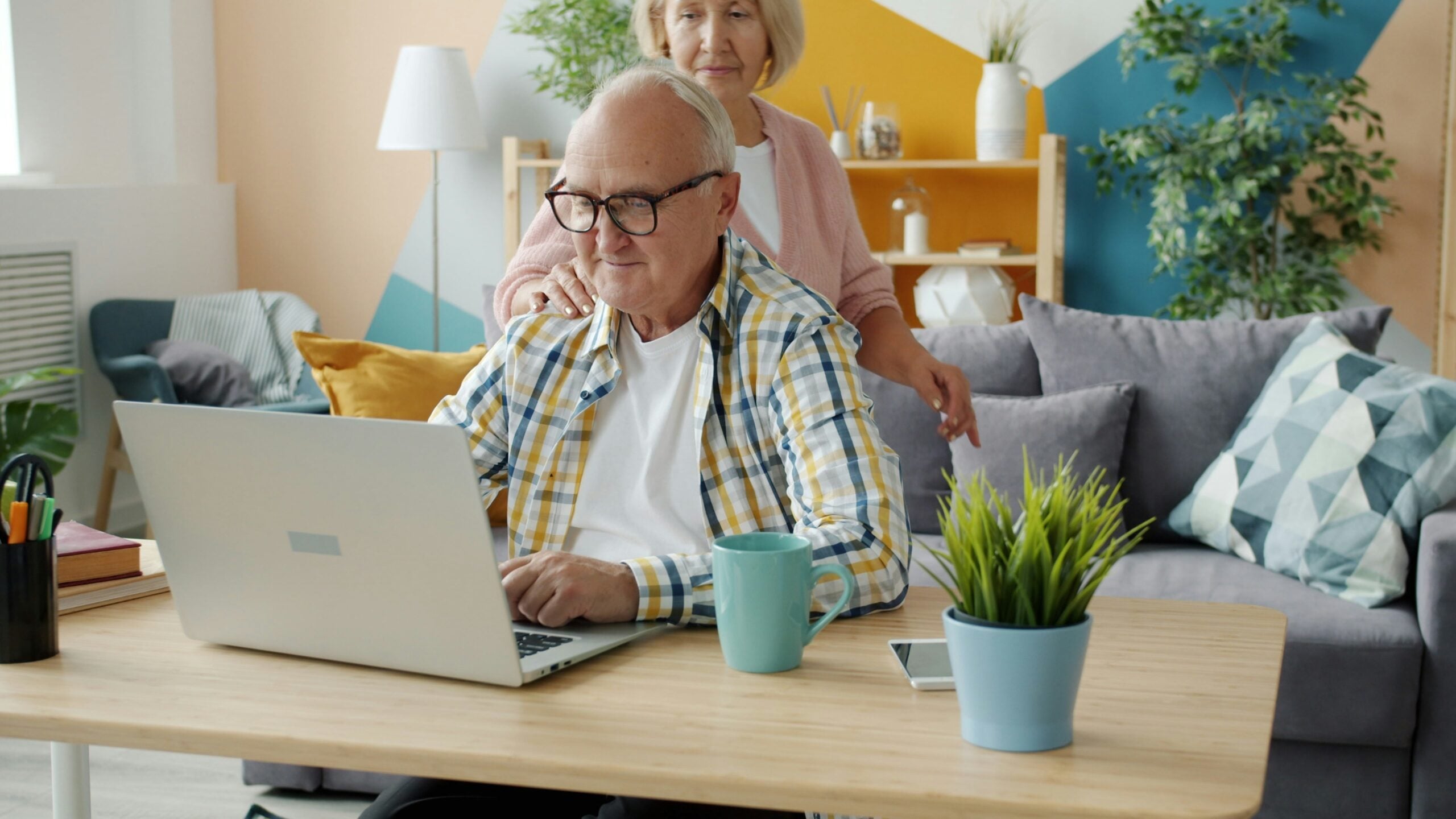 An older man uses a laptop at a table while a woman stands behind him, smiling, perhaps sharing some Boomer facts. The room is colorful with a couch, plants, and shelves in the background.