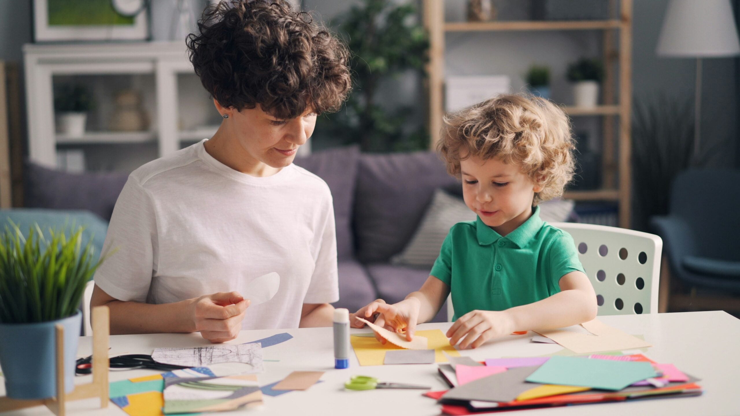 An adult and a child sit at a table doing arts and crafts with colorful paper, glue, and scissors in a cozy living room, capturing the simple joys of life in 2026.