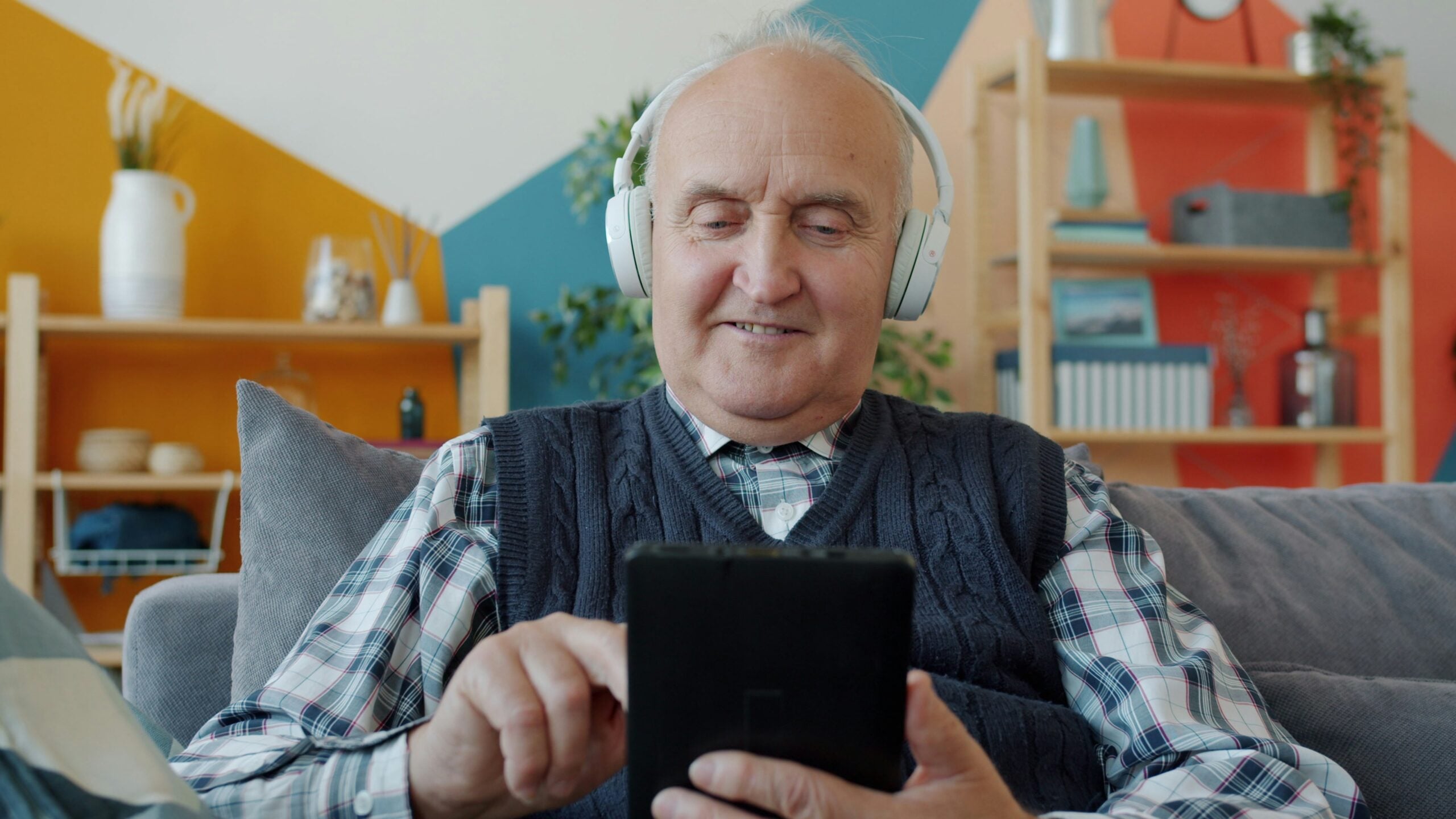 Older man wearing headphones smiles while using a tablet, sitting on a couch in a cozy, colorful living room with shelves and decorative items in the background—highlighting how boomers saving money can embrace technology for smarter financial choices.
