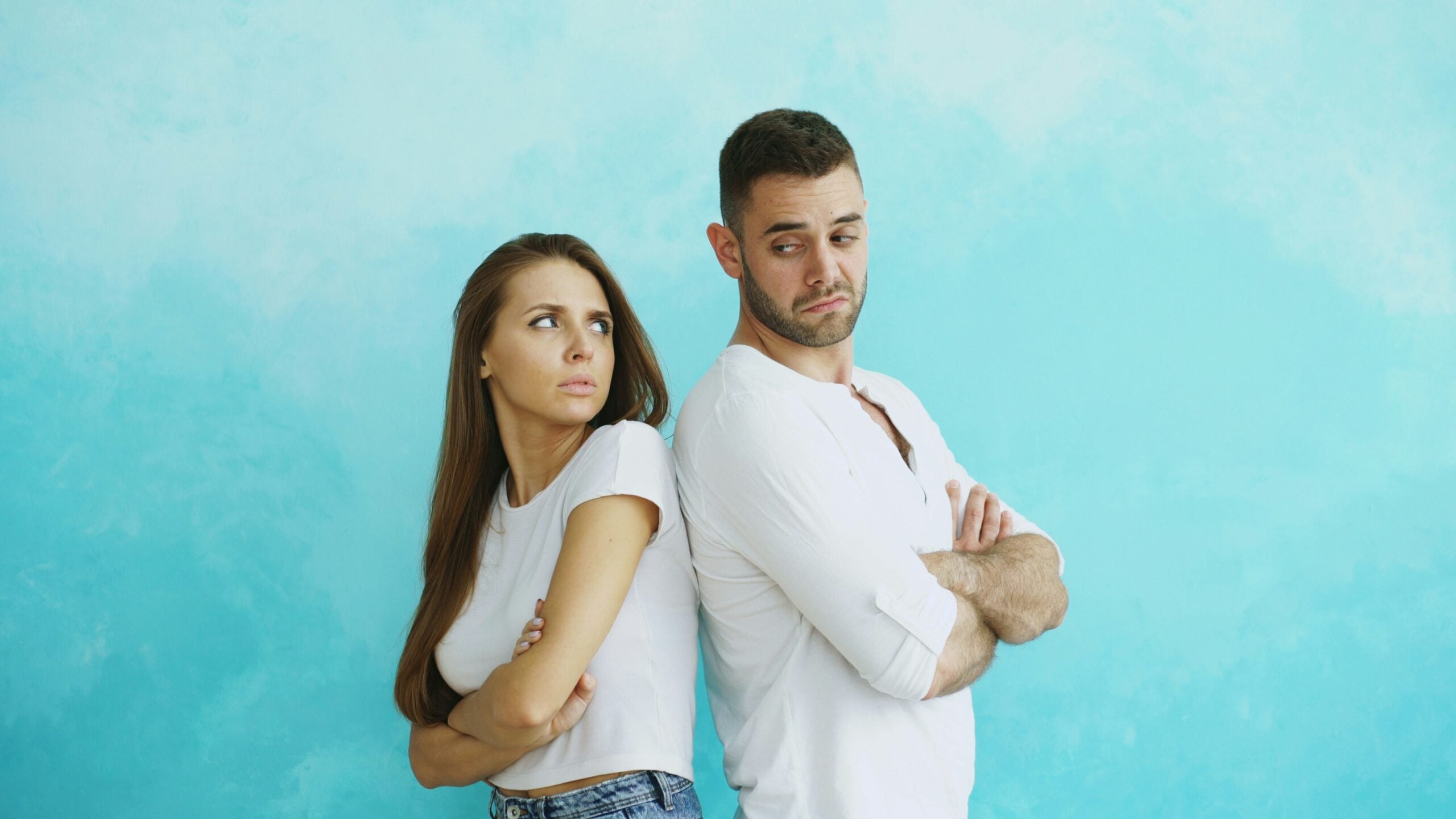 A woman and a man stand back-to-back with arms crossed, both looking at each other with skeptical expressions, perhaps contemplating the challenges of life in 2026 problems, against a blue background.