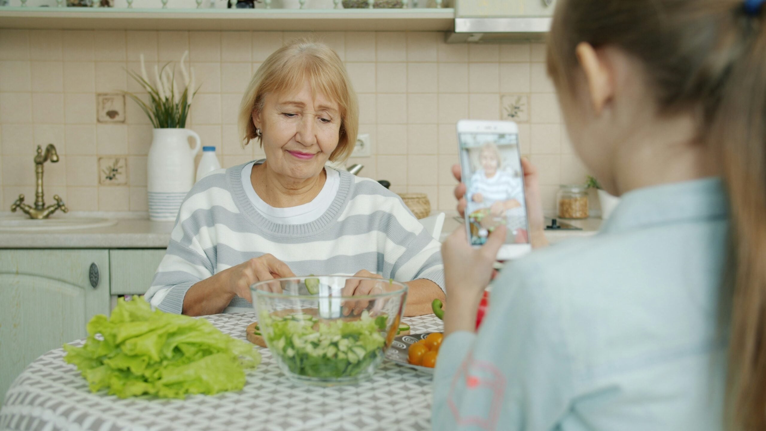 A young girl in the foreground holds up her smartphone to record a video of her grandmother, who sits at a kitchen table smiling while chopping cucumbers for a salad—a simple yet heartwarming glimpse into life in 2026. A bowl of fresh salad and lettuce are on the table.