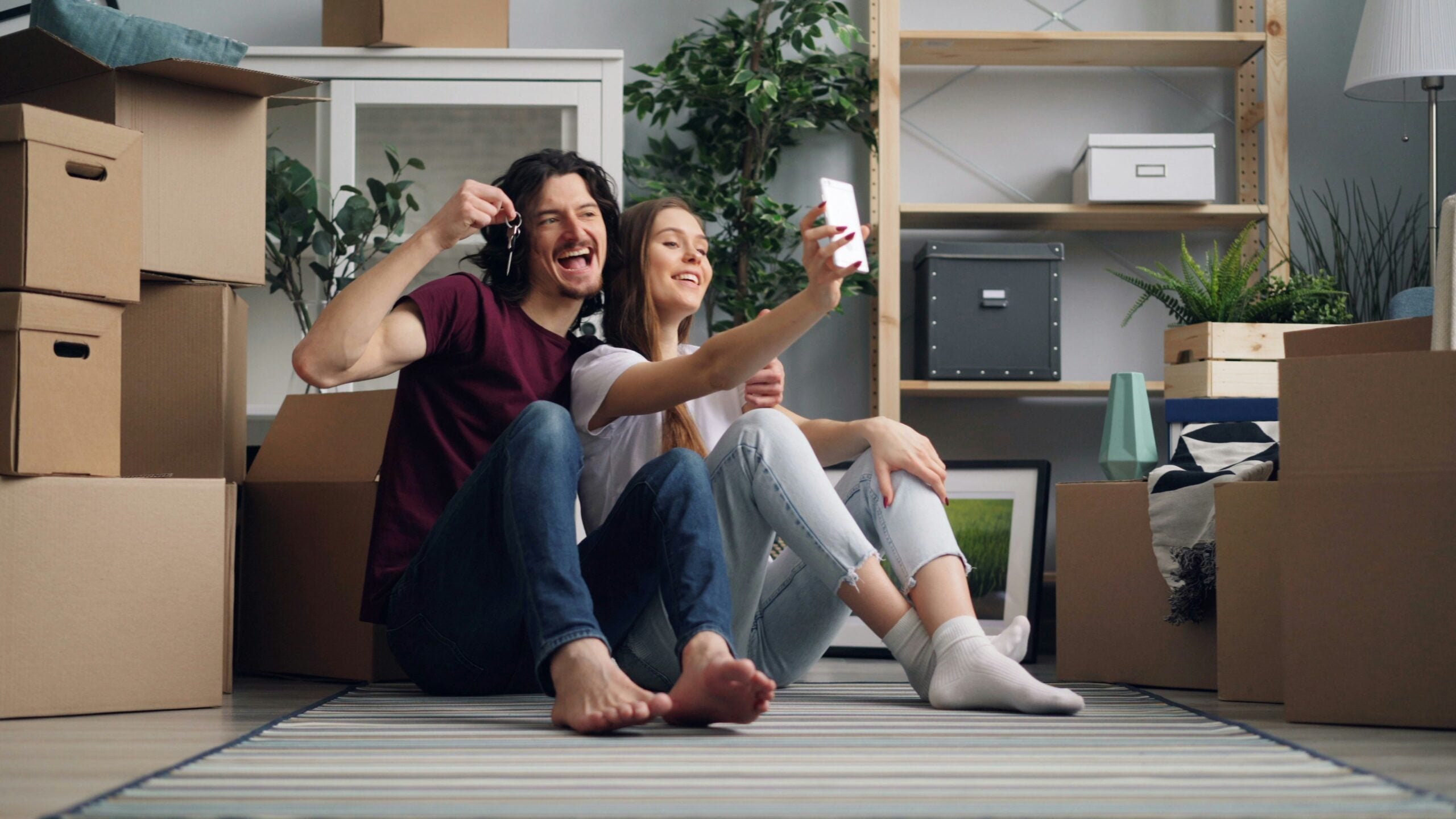 A smiling couple sits on the floor surrounded by moving boxes, taking a selfie. One person holds up keys, suggesting they’ve just moved into a new home—a perfect snapshot of life in 2026. Plants and shelves are visible in the background.