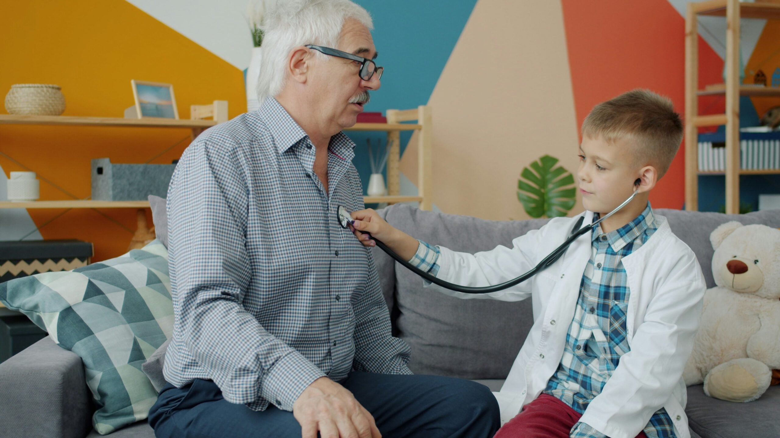 A young boy, dressed as a doctor, uses a stethoscope to listen to an older man’s chest in a colorful, cozy room with a teddy bear on the couch—perhaps getting ready to share some fun Boomer facts during their playful checkup.
