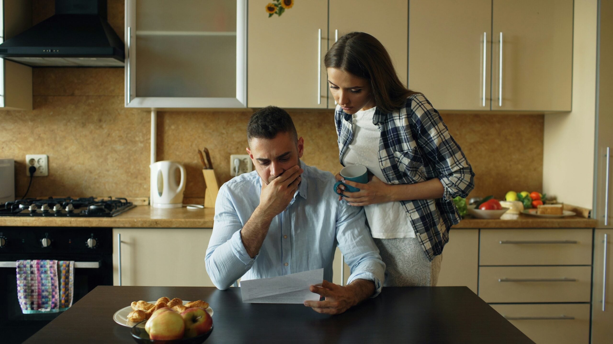 A man sits at a kitchen table looking worried while reading a paper, perhaps reflecting on the uncertainties of life in 2026. A woman stands beside him, holding a mug and looking concerned. Fruit and pastries are on the table.