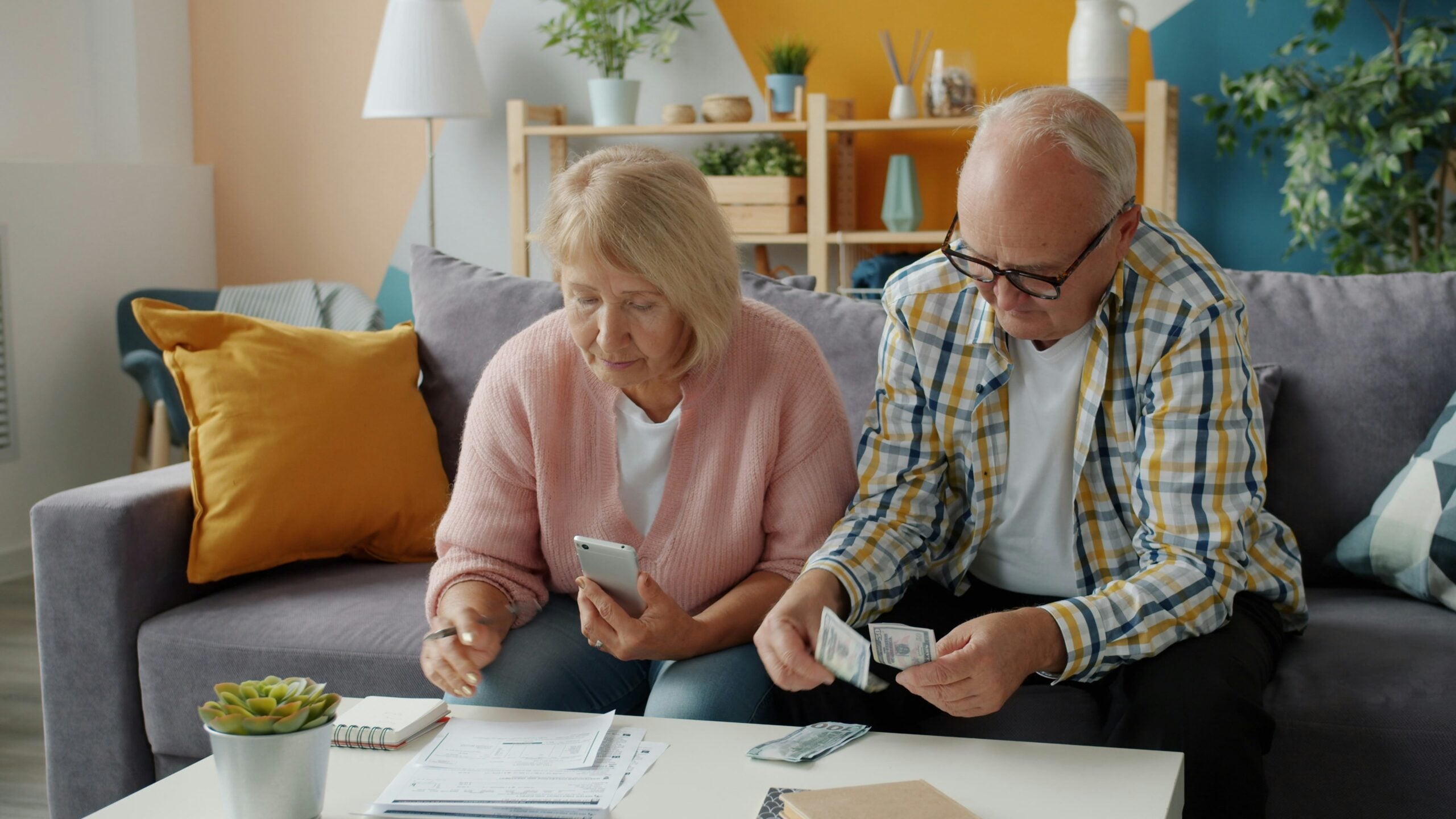 An older couple sits on a couch, reviewing bills and counting cash at a coffee table with papers, a calculator, and a phone, surrounded by boomer facts subtly displayed on the bookshelf in their cozy living room with yellow accents.