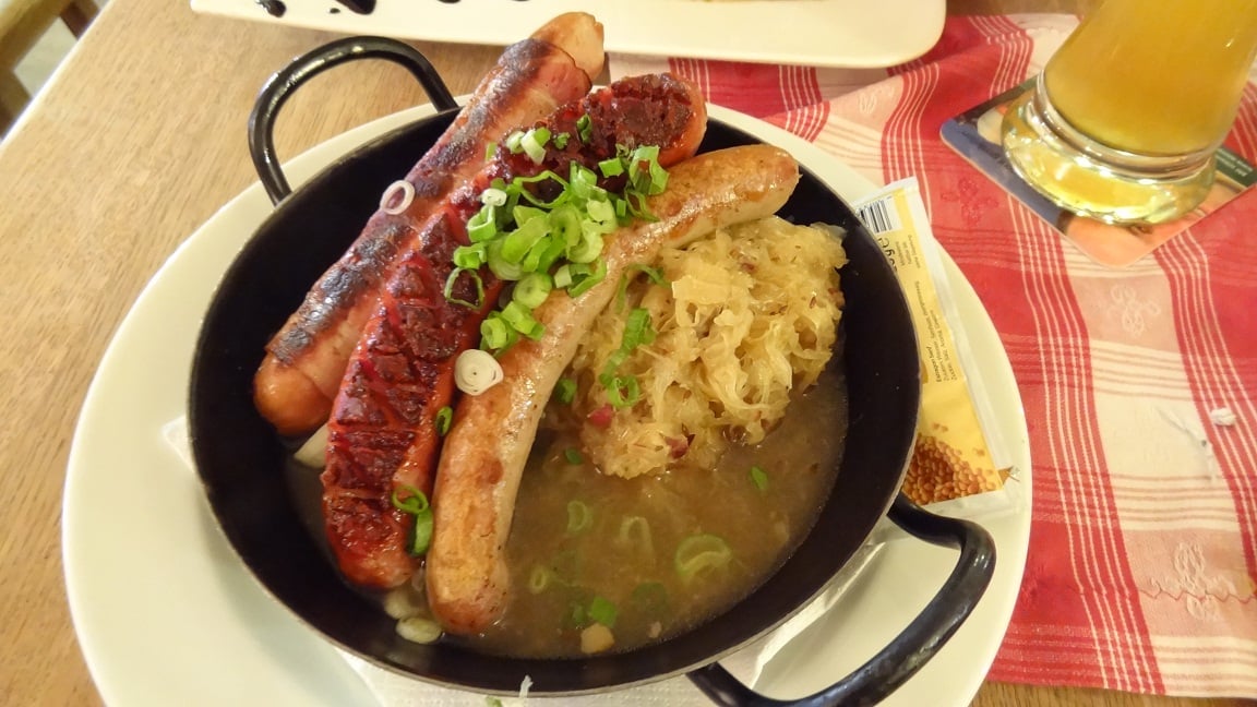 Three grilled sausages topped with chopped green onions served in a black bowl with sauerkraut and broth, on a white plate with a red checkered napkin and a glass of beer nearby—exactly the kind of amazing food people ate while on vacation.