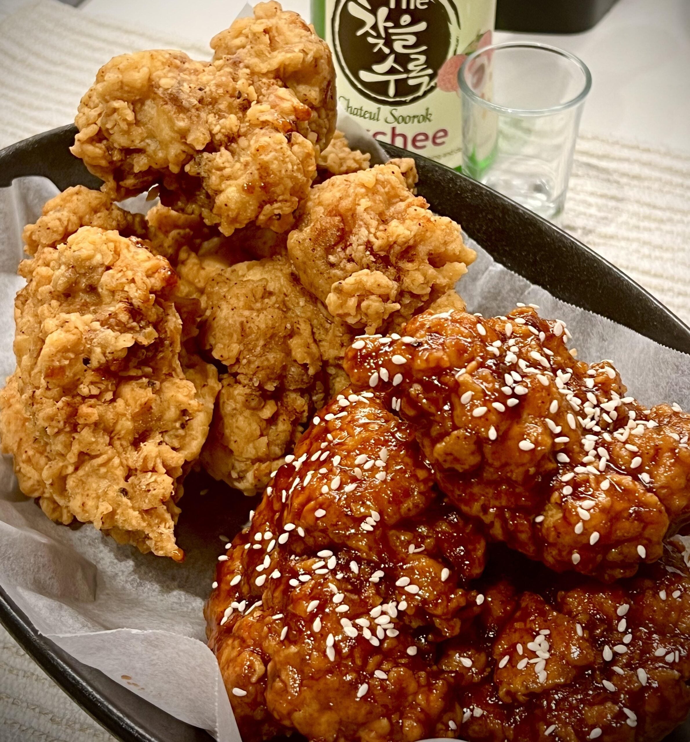 A plate with crispy fried chicken and saucy Korean fried chicken topped with sesame seeds, served on parchment paper with a bottle and shot glass in the background.