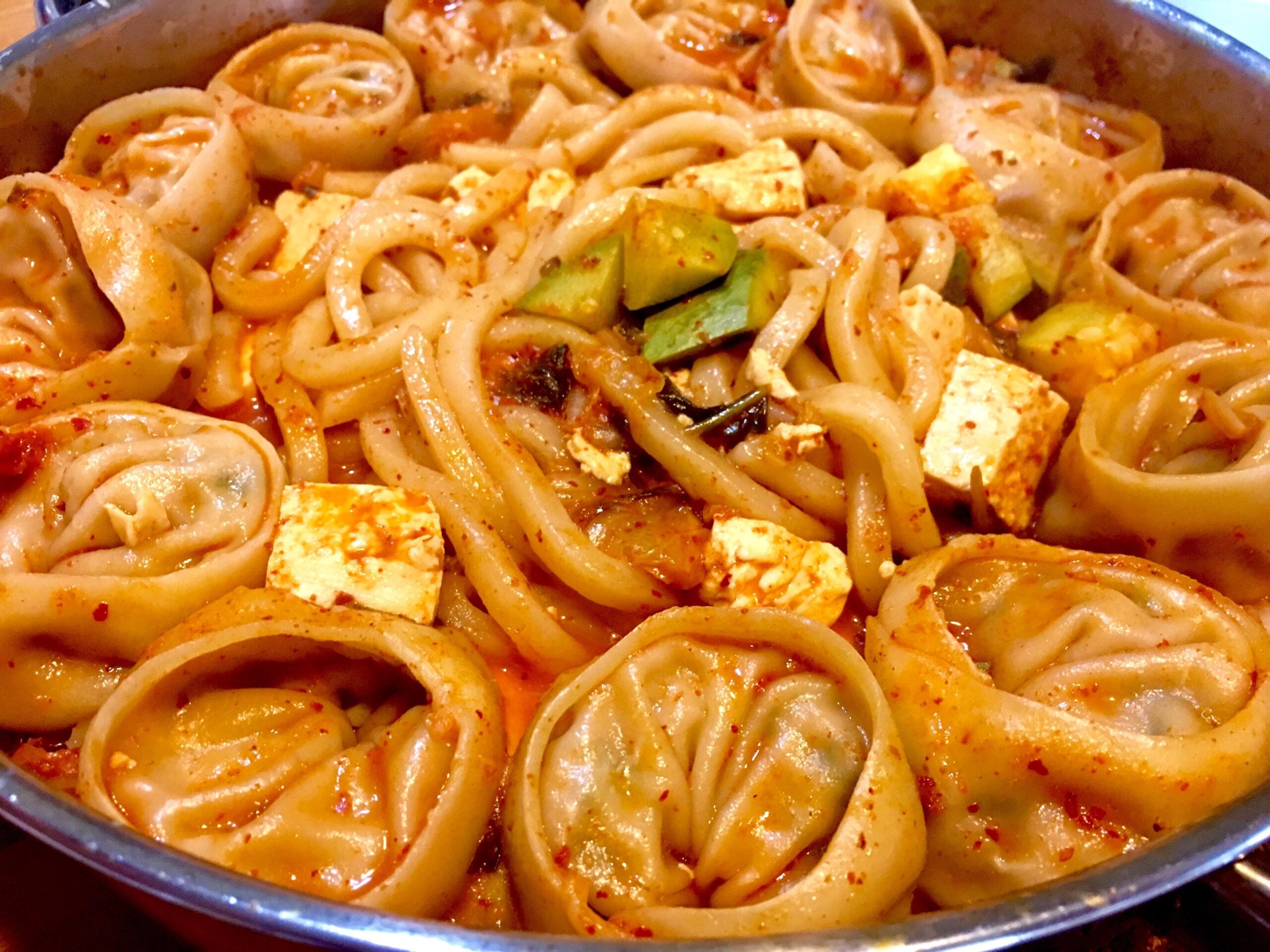 A close-up of a spicy Korean stew with round dumplings, thick noodles, tofu cubes, and vegetables in a red broth.