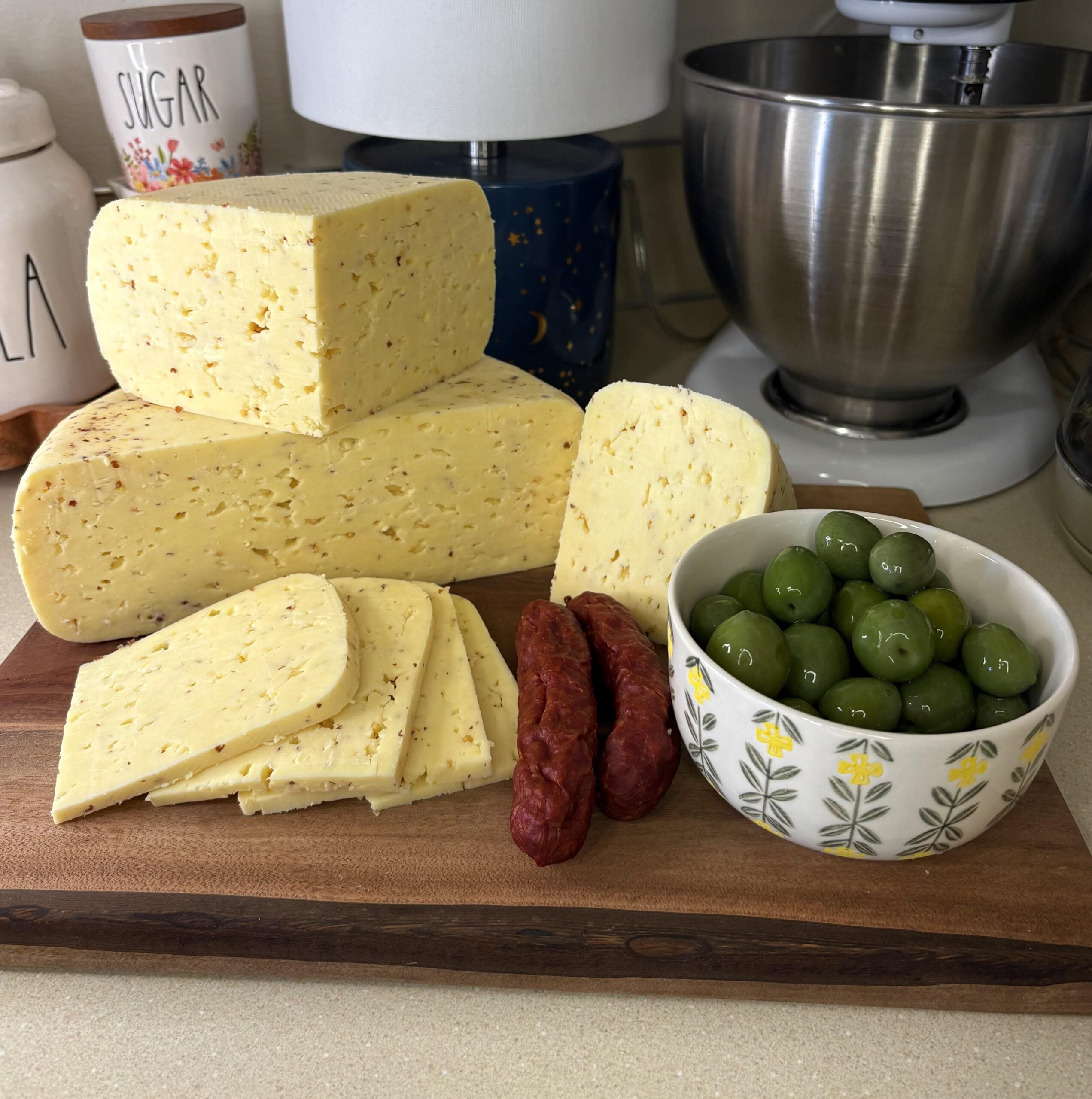 A wooden board with slices and blocks of cheese, two cured sausages, and a bowl of green olives. Kitchen items like a mixer, sugar container, and lamp are visible in the background.