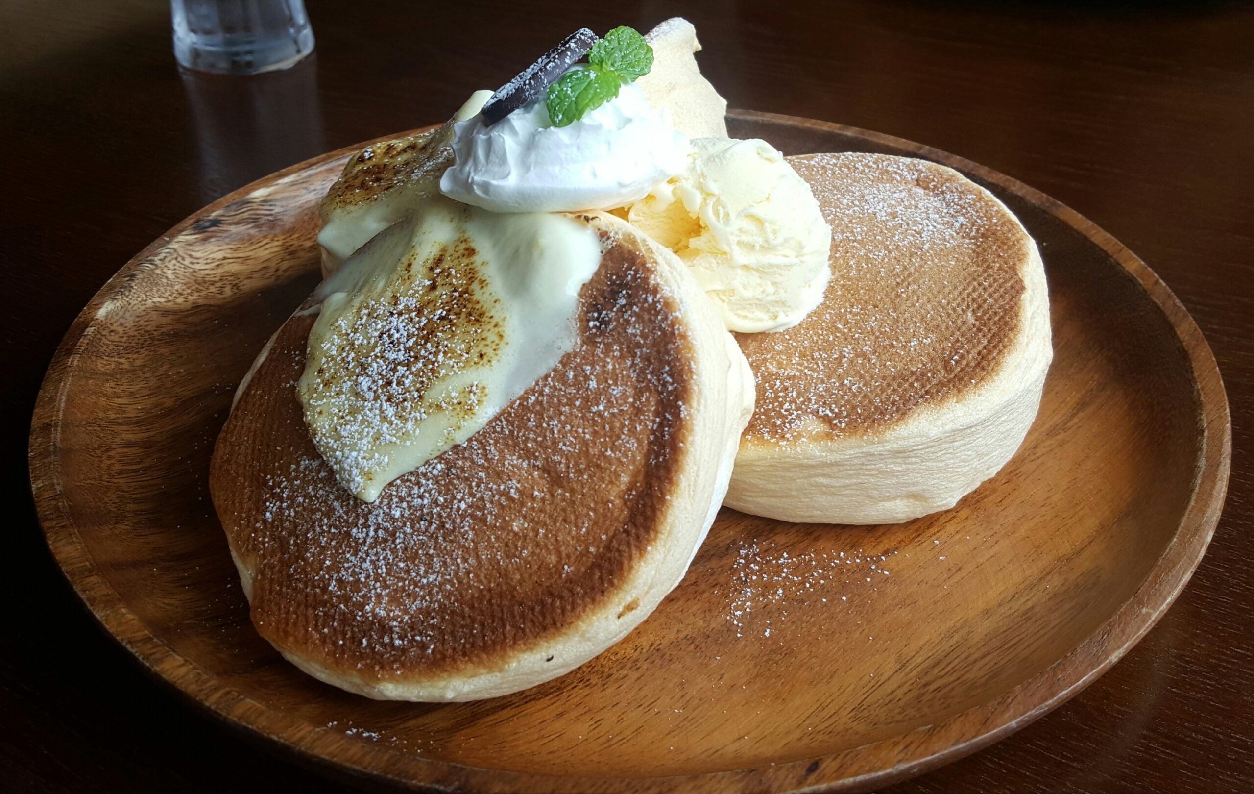 Two fluffy pancakes topped with whipped cream, a mint leaf, a chocolate piece, and a scoop of vanilla ice cream, served on a wooden plate and sprinkled with powdered sugar.