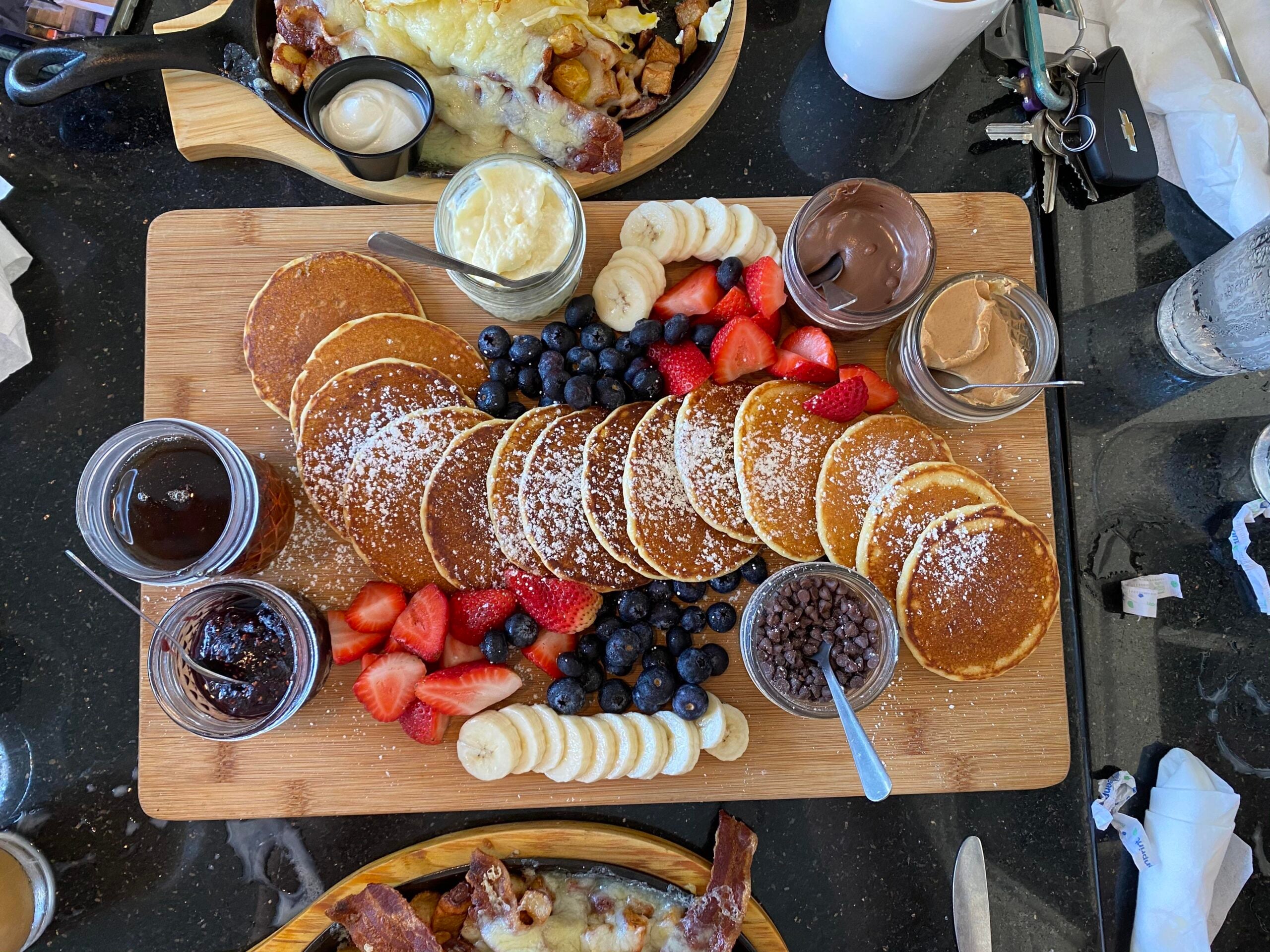 A wooden board with mini pancakes, banana slices, strawberries, blueberries, chocolate chips, and several jars of spreads, surrounded by plates of food and utensils on a black table.