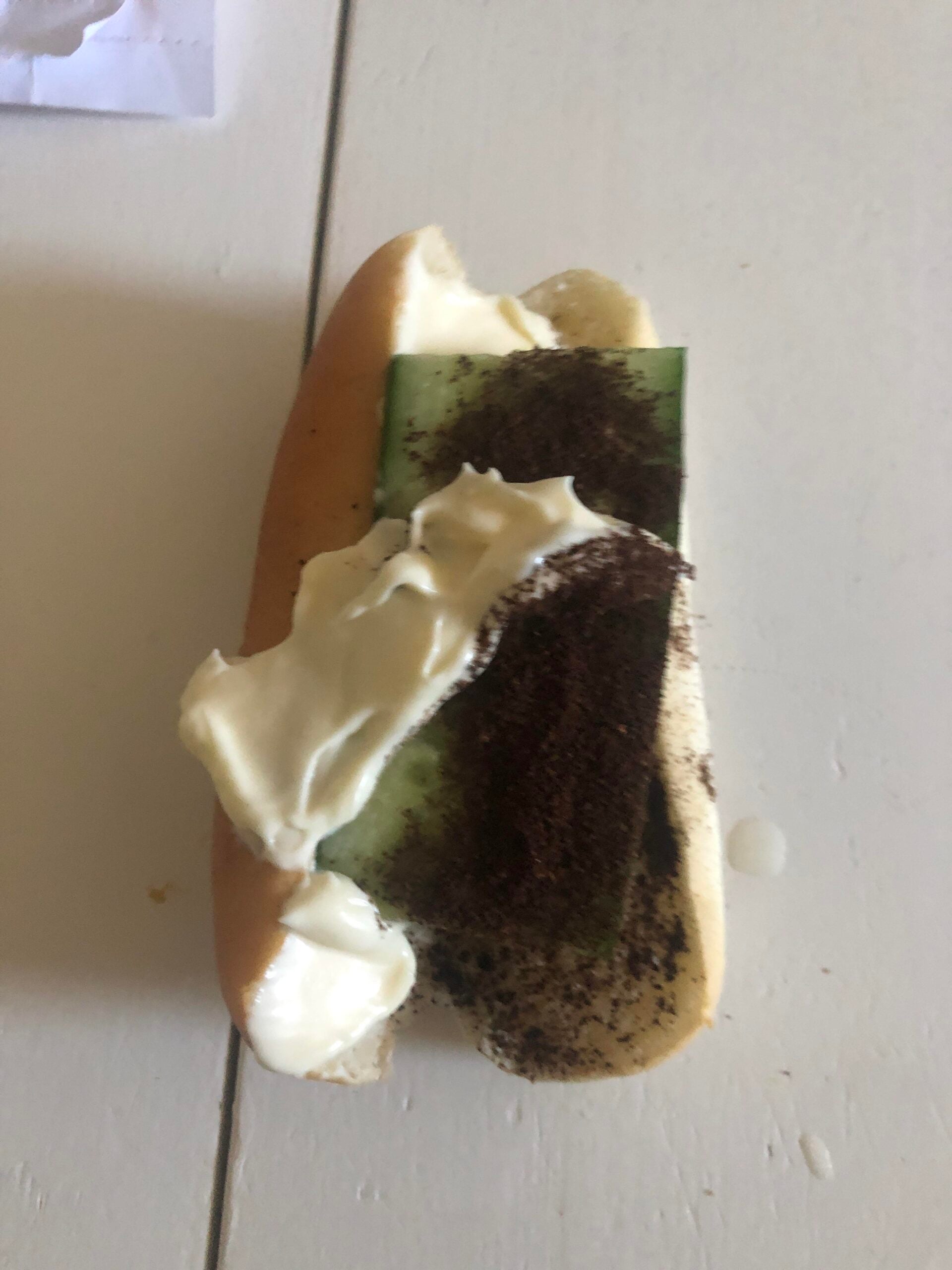 A hot dog bun filled with a slice of cucumber, topped with a generous amount of white cream and a sprinkle of dark powder, resting on a light-colored surface.