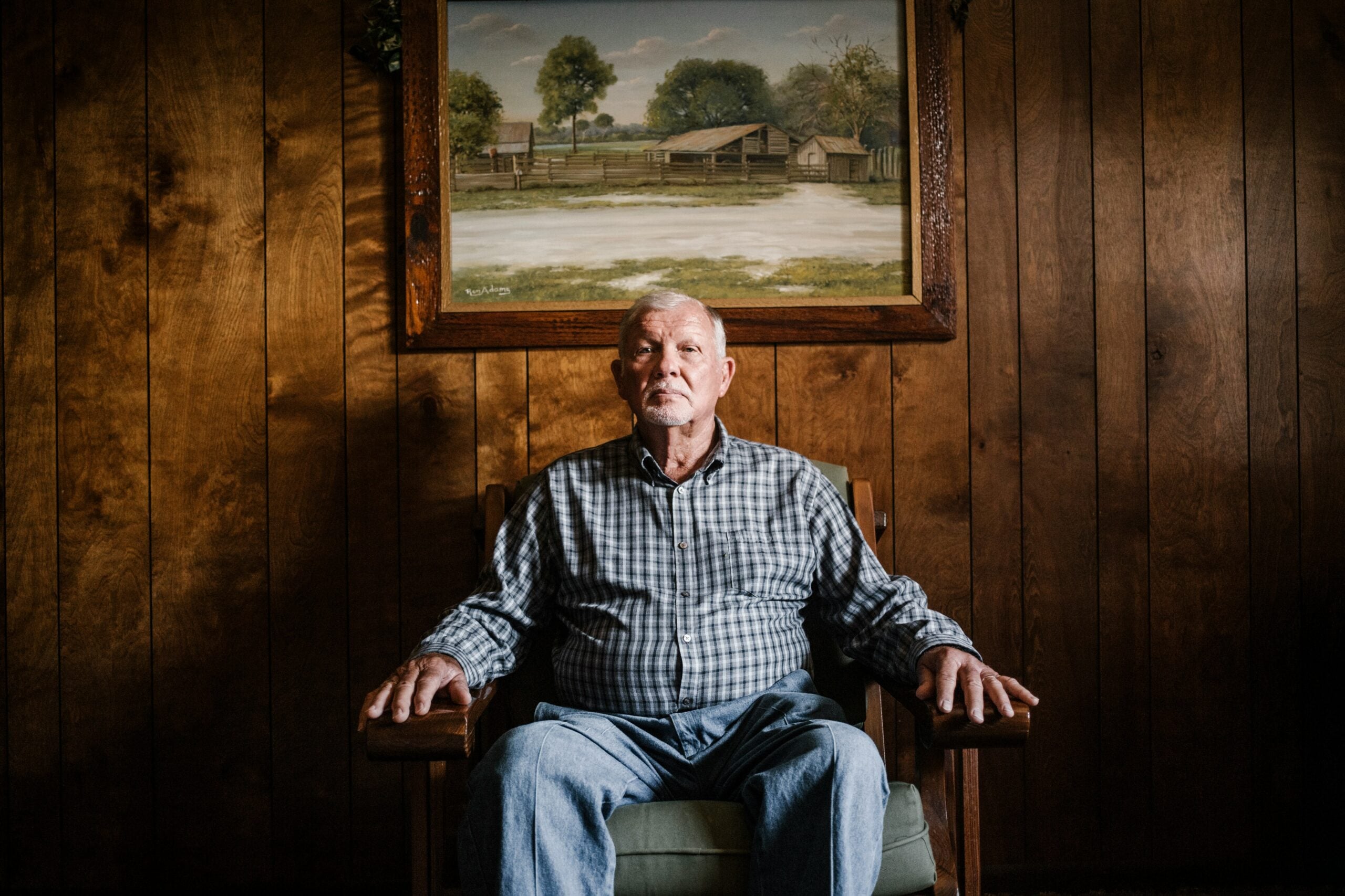 An older man sits in a wooden chair against a wood-paneled wall, his hands resting on the armrests, quietly reflecting on life in 2026. A framed painting of a rural scene hangs on the wall above and behind him.