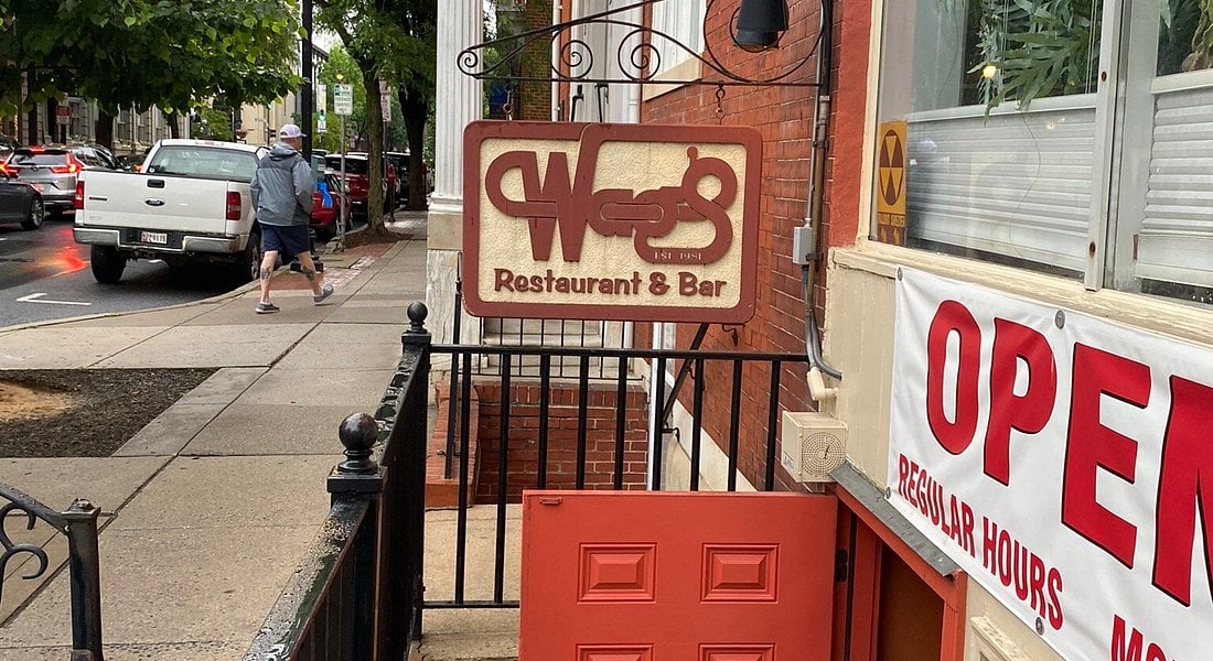 A brown and cream sign reading “West Restaurant & Bar” hangs above a red door on a city sidewalk. A banner with “OPEN” is visible, and a person walks by on the street.