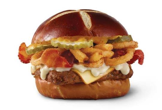 A sandwich with a pretzel bun, fried chicken, white cheese, crispy onion rings, pickles, bacon, and sauce, against a plain white background.